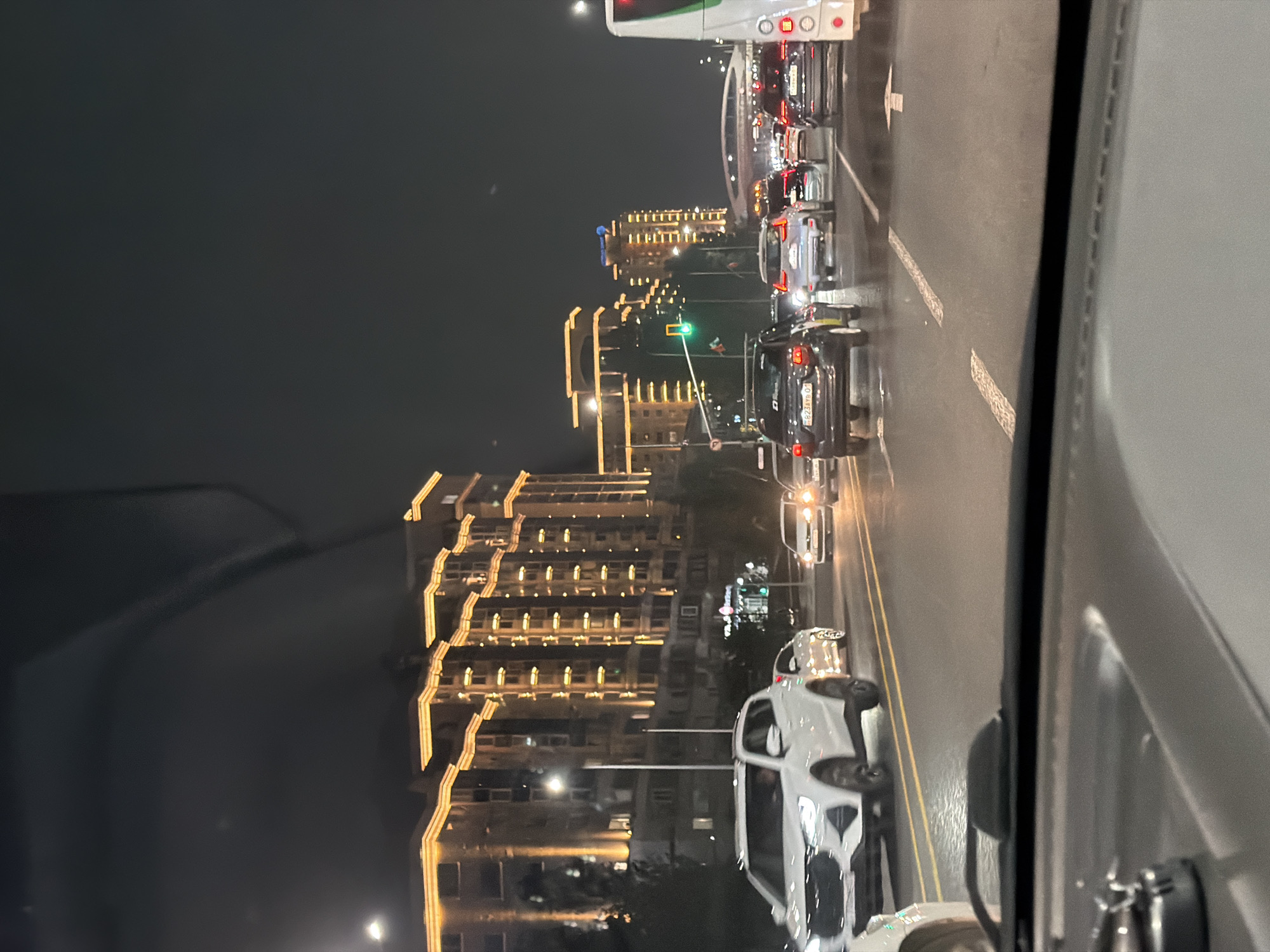 The image depicts a nighttime street scene in Astana, Kazakhstan, likely along a major avenue such as one of the main roads leading to or from the city center, characterized by modern high-rise buildings illuminated with decorative lights. In the foreground, the dashboard of a vehicle is visible, suggesting the photo was taken from inside a car. The road ahead is filled with traffic, with numerous cars, SUVs, and a bus visible. Vehicles are traveling in multiple lanes, and traffic lights are present, with one appearing green. The buildings lining the street are tall, multi-story structures with illuminated facades, showcasing a contemporary urban landscape. The overall atmosphere is one of a busy city at night, with artificial lights reflecting off the wet road surface, indicating recent rain or a damp environment.