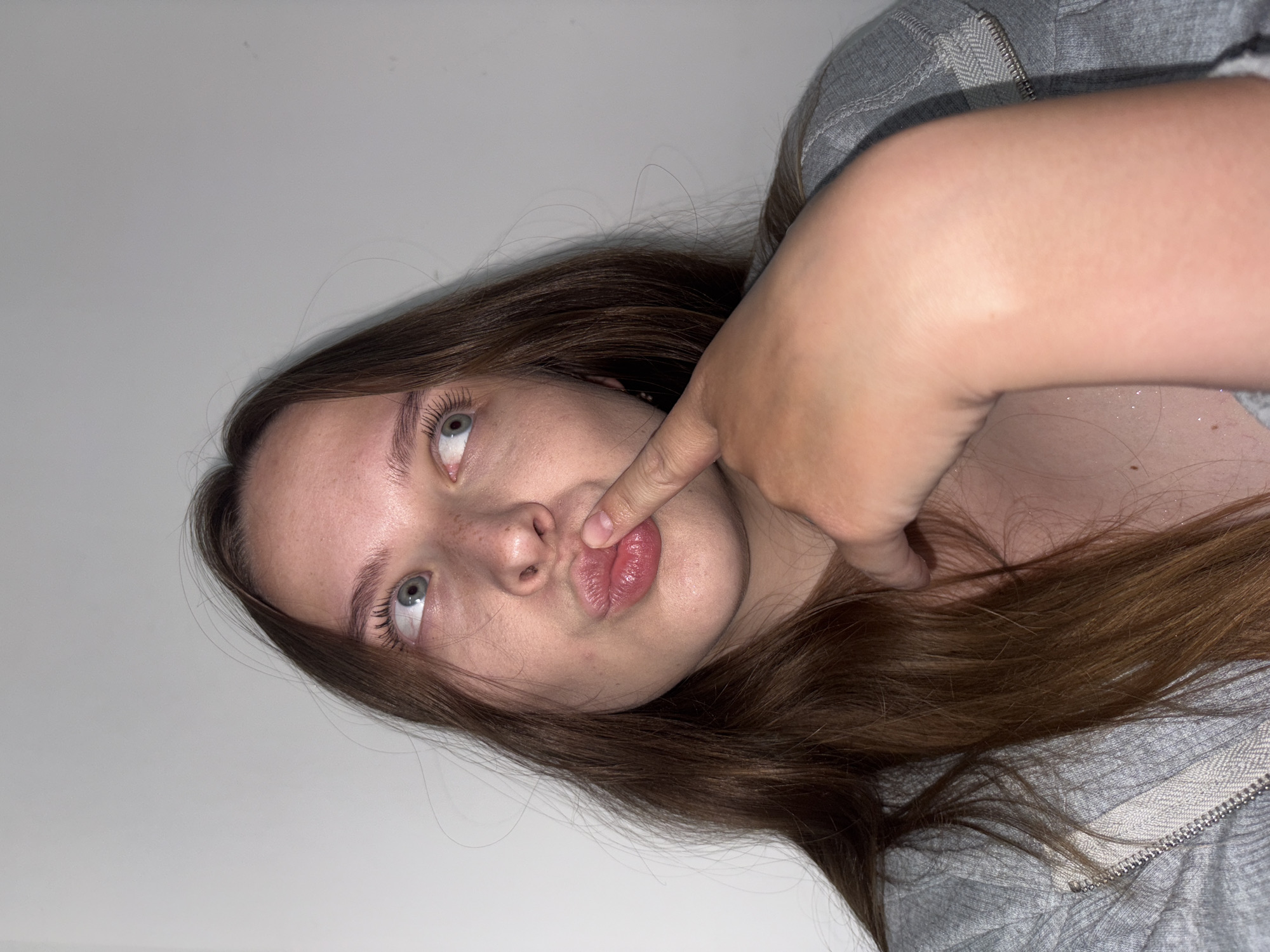 A young woman with long, brown hair and blue eyes is looking to her right, with her finger touching her lips. She has freckles on her nose and cheeks. She is wearing a gray zip-up hoodie. The background is a plain white wall. The image does not provide enough information to determine its location in Chaikovsky, Russia.