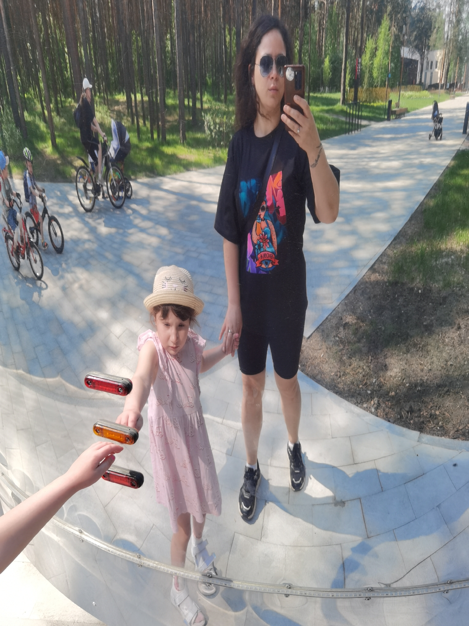 The image captures a woman taking a selfie with a young girl in a park-like setting, reflected in a curved, mirrored surface. The location is Perm, Russia.
The woman is wearing sunglasses, a dark t-shirt with a colorful graphic, and dark shorts. The young girl is wearing a light pink dress and a straw hat. They are holding hands, standing close to the mirrored surface.
In the reflection, the background shows a paved path, trees, and other people. Some people are riding bicycles and others are walking. The mirrored surface shows additional reflections, including a child's hand reaching towards the glass, positioned near the image of the girl, and several orange and red reflectors are visible in the reflection.