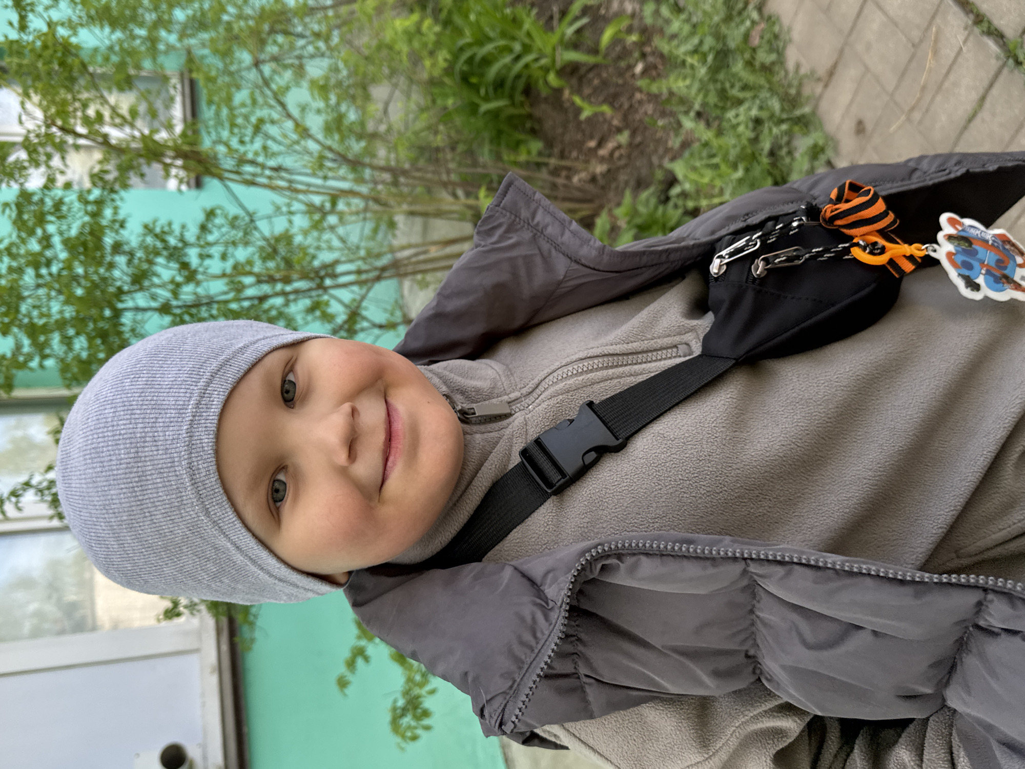 A child wearing a grey beanie, a grey fleece, and a grey puffy jacket poses outdoors. They are wearing a black fanny pack with a striped orange and black ribbon and a cartoon keychain attached. Behind the child are green bushes, a light blue building, and a sidewalk. The location is Chaikovsky, Russia.