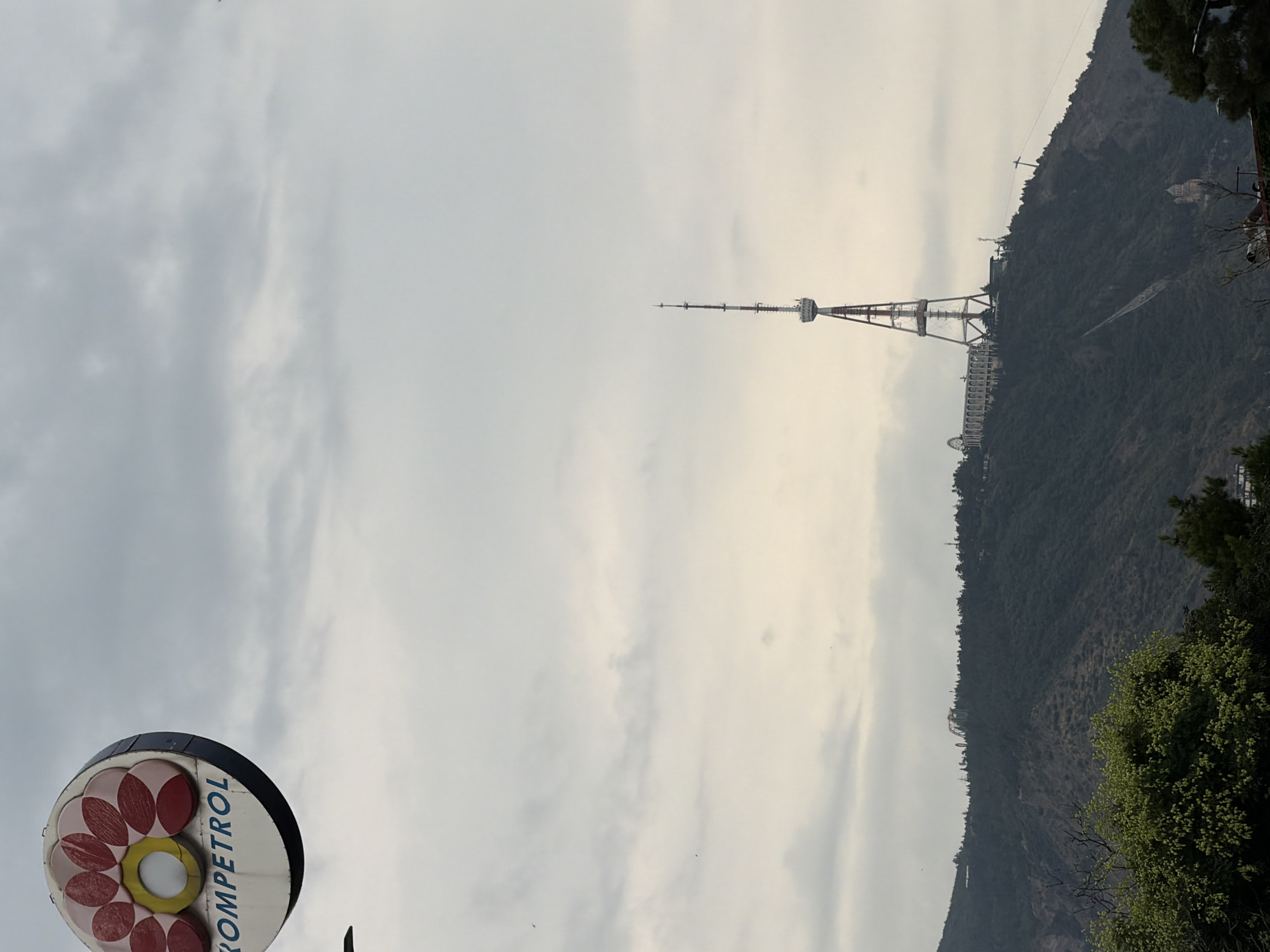 The image depicts a wide shot of the Tbilisi TV Tower and the surrounding hillside, with a sign for "KOMPETROL" visible in the upper left corner. The sky is overcast, with a soft, diffused light. The TV tower, a prominent landmark in Tbilisi, is located on top of a heavily forested hill, with some buildings visible at its base. The "KOMPETROL" sign, circular in shape with a flower-like design, is also mounted on a pole and appears to be advertising a company or service. The overall impression is one of a cloudy day in a mountainous urban environment.