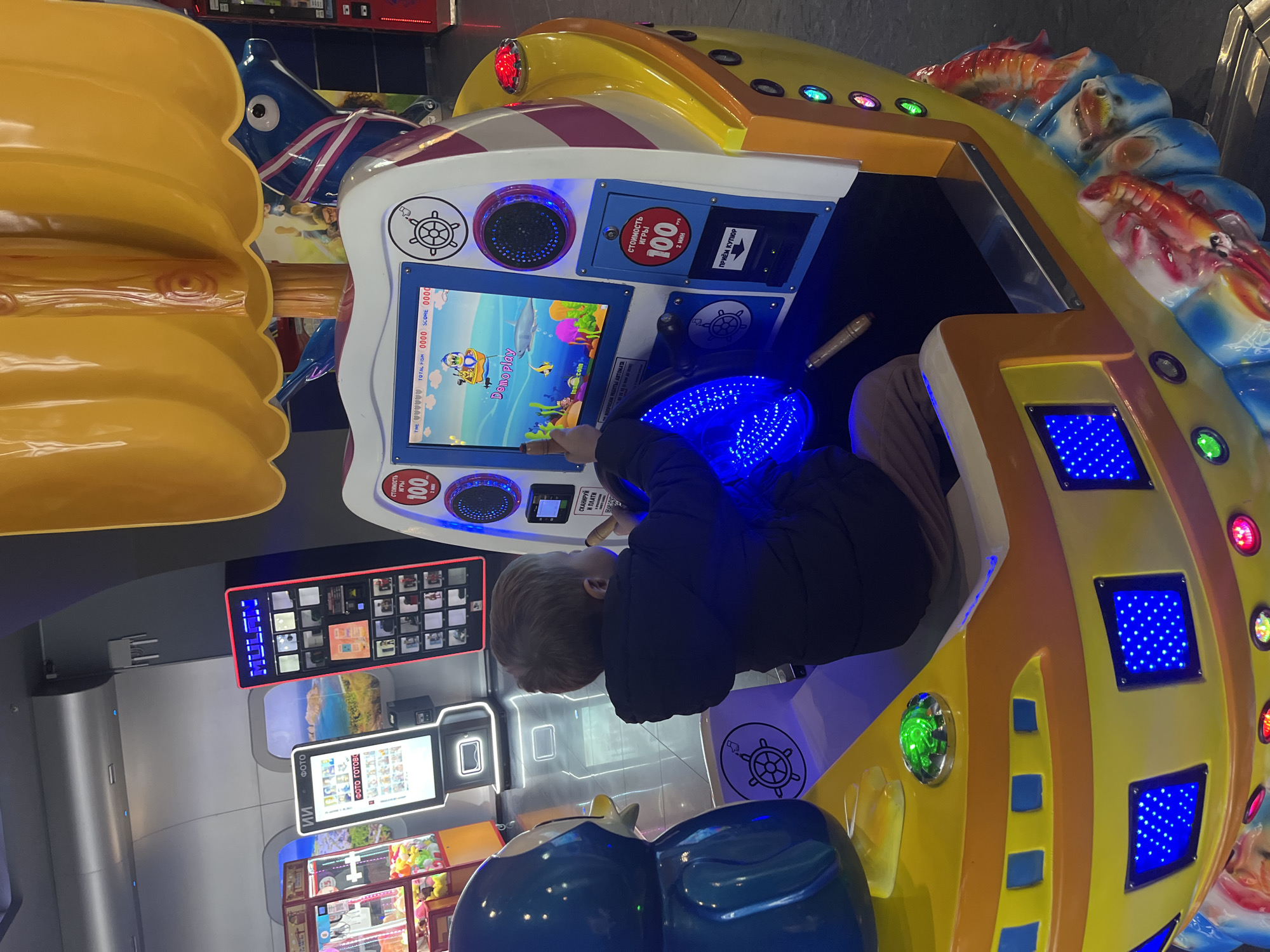 A young child, wearing a dark blue puffer jacket and light brown pants, is intensely focused on playing an arcade game. The child, seen from behind, has light brown hair and is actively engaging with the game's controls, with one hand touching the screen and the other near a brightly lit blue steering wheel or joystick.
The game machine is shaped like a colorful boat, predominantly yellow and white with blue and red accents. It features decorative elements such as stylized fish and shrimp along its top edge. Blue LED lights illuminate parts of the machine, including a small panel and the steering wheel area. The game screen displays a cartoon-like underwater scene with a character, fish, and text indicating "Demoplay" and scores ("TOTAL FISH 0000 SCORE 0000").
Visible Russian text on the game machine includes "стоимость игры 100" (cost of game 100), "2 мин" (2 min), and "приём купюр" (accepts banknotes), indicating the game costs 100 units of currency (likely Russian Rubles) for a 2-minute play. Another sign says "СКАНИРУЙ И ПЛАТИ" (Scan and Pay).
In the background, other arcade machines or photo booths are visible. One machine displays "ФОТО" (Photo) and "ФОТО ГОТОВО" (Photo ready) in Russian. The setting appears to be an indoor amusement center or arcade, characterized by artificial lighting and multiple electronic game stations. The child appears to be completely engrossed in the game, demonstrating concentration and engagement.