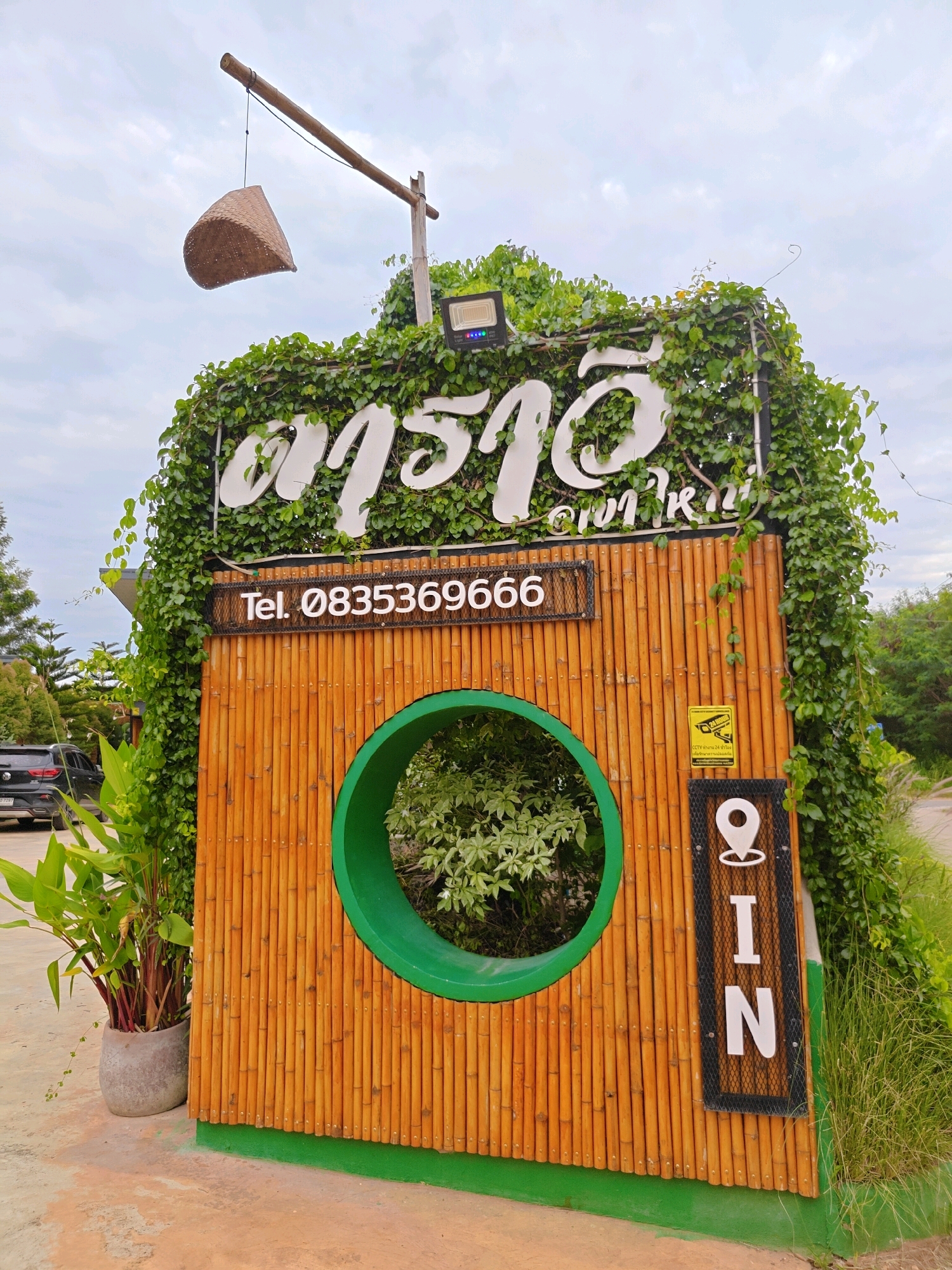 The image shows a facade made of bamboo poles, overgrown with green vines. A large, white, ornate sign with Thai lettering is prominently displayed at the top. Below the Thai sign, a smaller black sign with white lettering reads "Tel. 08353696966". Centered on the bamboo facade is a large, circular opening with a green border, revealing greenery within. To the right of this opening, a vertical black sign with white lettering spells out "G I N", with a white location pin icon at the top. Below the "G I N" sign, a yellow security camera sticker is affixed to the bamboo. A portion of a black car and some green plants are visible to the left. The overall setting appears to be outdoors, likely in a tropical or subtropical environment, given the vegetation. The cloudy sky suggests it is daytime. The scene is located in Mu Si, Thailand.