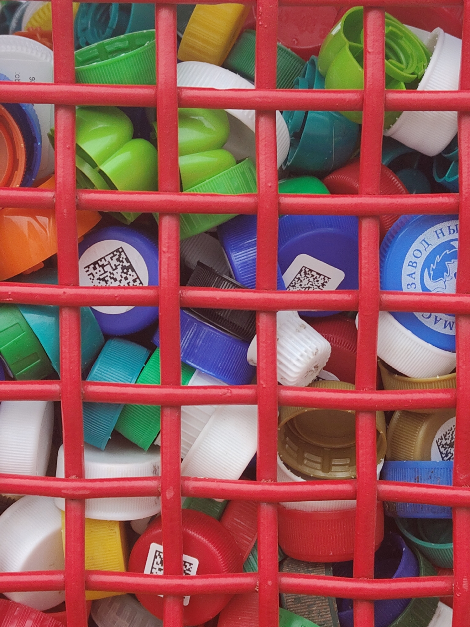 A high-angle, close-up shot shows a red, metal grid enclosing a collection of colorful plastic bottle caps. The caps are predominantly green, white, blue, red, and yellow. Some caps have printed labels or QR codes. The scene's location is an unknown city, in an unknown country.