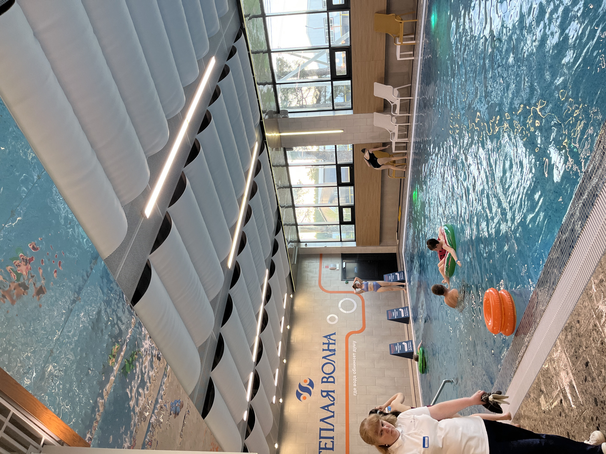 The image depicts an indoor swimming pool facility, likely located in Votkinsk, Russia, based on the Cyrillic text on the wall ("ТЕПЛАЯ ВОЛНА," which translates to "WARM WAVE"). The pool is filled with blue water, with people swimming and using inflatable rings. A woman in a white shirt stands near the edge of the pool, potentially an employee. Diving blocks are positioned along one side of the pool. The facility features a high ceiling with a distinct wave-like pattern and integrated lighting. Windows are visible along one wall, providing natural light. Chairs are placed near the windows. A person is standing on the side of the pool facing the water. The overall scene suggests a leisure or recreational setting.