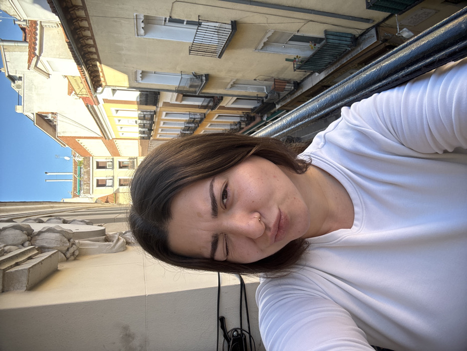 The image is a selfie taken by a young adult female on a balcony overlooking a narrow urban street in Madrid, Spain, under a clear blue sky. The female, centered in the mid-foreground, has shoulder-length dark brown hair and is wearing a plain white long-sleeved shirt. She has a small gold hoop piercing in her right nostril. Her expression is a playful wink, with her left eye closed and her lips pursed in a slight pout.
To the immediate left of the female, the ornate, cream-colored facade of the building she is on features detailed architectural bas-relief elements and a black cable running vertically. The lower right foreground shows a dark metal balcony railing.
The background reveals a densely built-up urban environment typical of Madrid. Below and across from the balcony, a narrow street is flanked by multi-story residential buildings. These structures display warm yellow, cream, and terracotta-colored facades, numerous windows, and dark-colored wrought-iron balconies. Some distant rooftops feature traditional red tiles and modern chimneys or vents. The scene is brightly illuminated by strong sunlight, indicating daytime conditions.