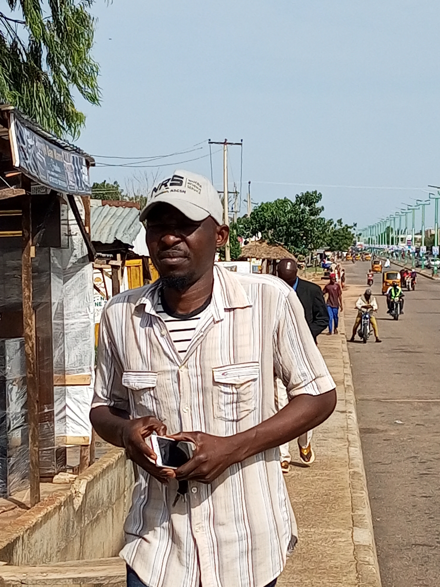 The image depicts a street scene in Tempure, Nigeria. In the foreground, a man wearing a striped shirt and a baseball cap stands on a sidewalk, holding a smartphone. He has a beard and appears to be looking towards the viewer. Behind him, other individuals and a variety of vehicles, including motorcycles and cars, are visible on the road. The environment suggests a moderately busy urban area with some market stalls and vegetation present. The sky is clear and blue.