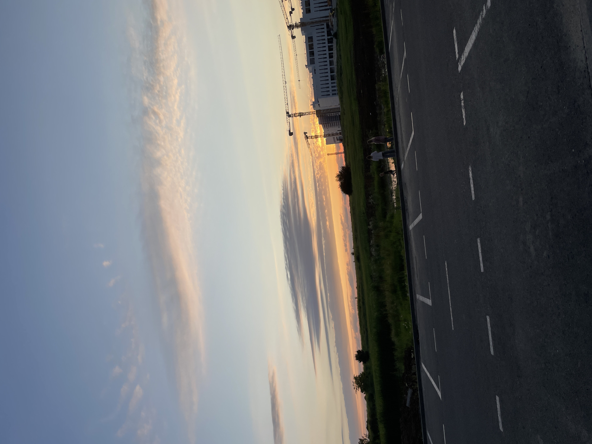 The image depicts a sunset scene with a sky filled with orange, pink, and blue hues. In the background, a large building complex is under construction, with several cranes visible. A grassy field with some trees is located between the building and a body of water. In the foreground, there is an asphalt parking lot with white markings. Three people, appearing to be adults and a child, are standing near the water's edge, facing the sunset. The scene appears to be a suburban or outskirts area of Stavropol, Russia, given the combination of construction and open land.