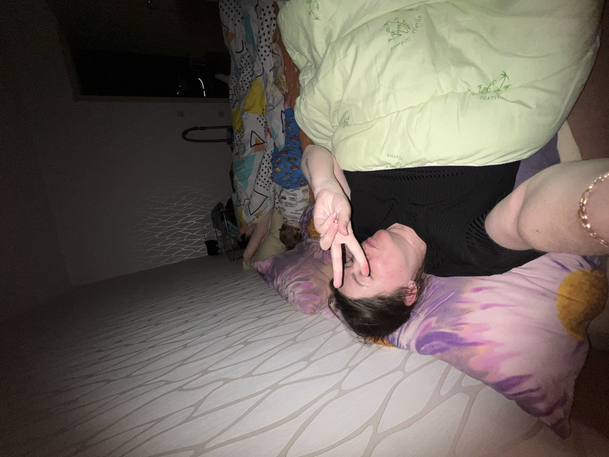 The image captures an indoor scene, likely a bedroom, characterized by low light, suggesting nighttime or a dimly lit environment. The primary subject is a woman lying on a bed, resting her head on a vibrant tie-dye pillow featuring purple, pink, and yellow hues. She has dark hair and is wearing a dark top. With her right hand, she makes a peace sign, holding her fingers near her face, which is slightly tilted. A gold-colored chain bracelet is visible on her left wrist.
Behind the woman, partially obscured by bedding, another person, likely a child, is visible, covered by a colorful, patterned blanket. This blanket features a mix of white with black dots, yellow sections, and geometric shapes, with "PALINDRA" faintly discernible on it. The bed itself is covered by a light green comforter or duvet, which bears "BAMBOO TEXTILE" branding along with small bamboo tree logos. The mattress underneath has a subtle light gray wavy pattern.
The room's left wall and floor are visible, displaying faint, wavy patterns created by light reflections, adding to the atmospheric dimness. A dark, rectangular opening or window is in the upper left corner, and a metal structure, possibly a clothing rack or part of a bed frame, is nearby. Some indistinct objects and clutter are visible near the child. The overall mood appears relaxed and informal. While no specific visual cues confirm the location, the provided context indicates the image was taken in Perm, Russia.