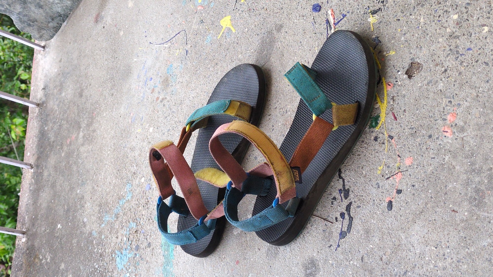 Two Teva sandals are lying on a speckled concrete surface. The sandal on the right has straps that are brown, green, and yellow. The sandal on the left has pink, green, and yellow straps. Splatters of various colors are on the concrete surface. A partial view of a railing is visible in the upper-left corner of the image, with green foliage behind it. The location is not evident.