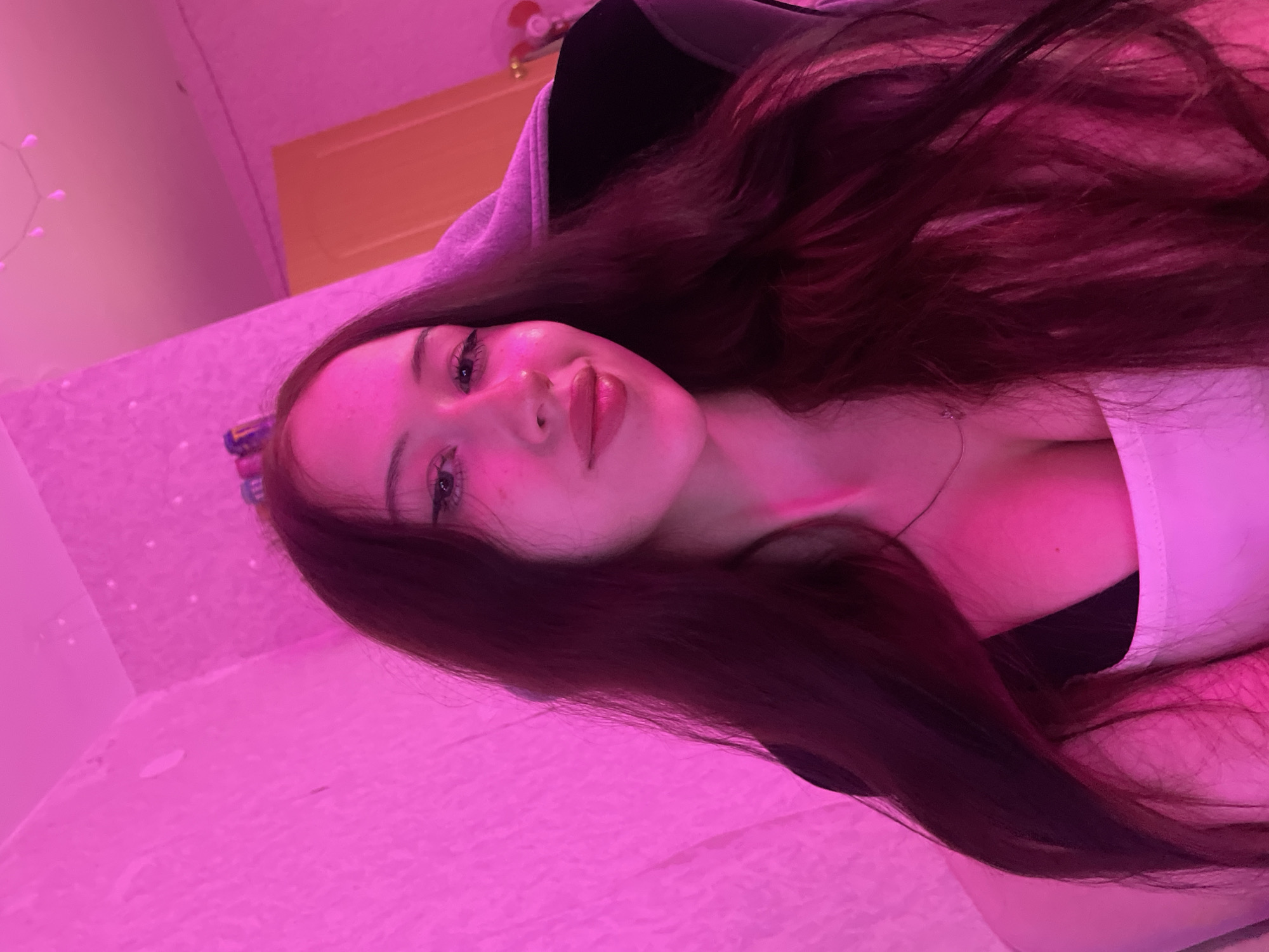 The image is a selfie of a young woman with long, dark hair and fair skin, illuminated by pink lighting. She is wearing a white top and a dark bra. Her makeup includes winged eyeliner and lipstick. In the background, there is a wall with a textured surface and a doorway. A desk chair is visible behind her. The setting appears to be an indoor residential space, possibly a bedroom or gaming room. There is no specific information to place this image in Votkinsk, Russia.