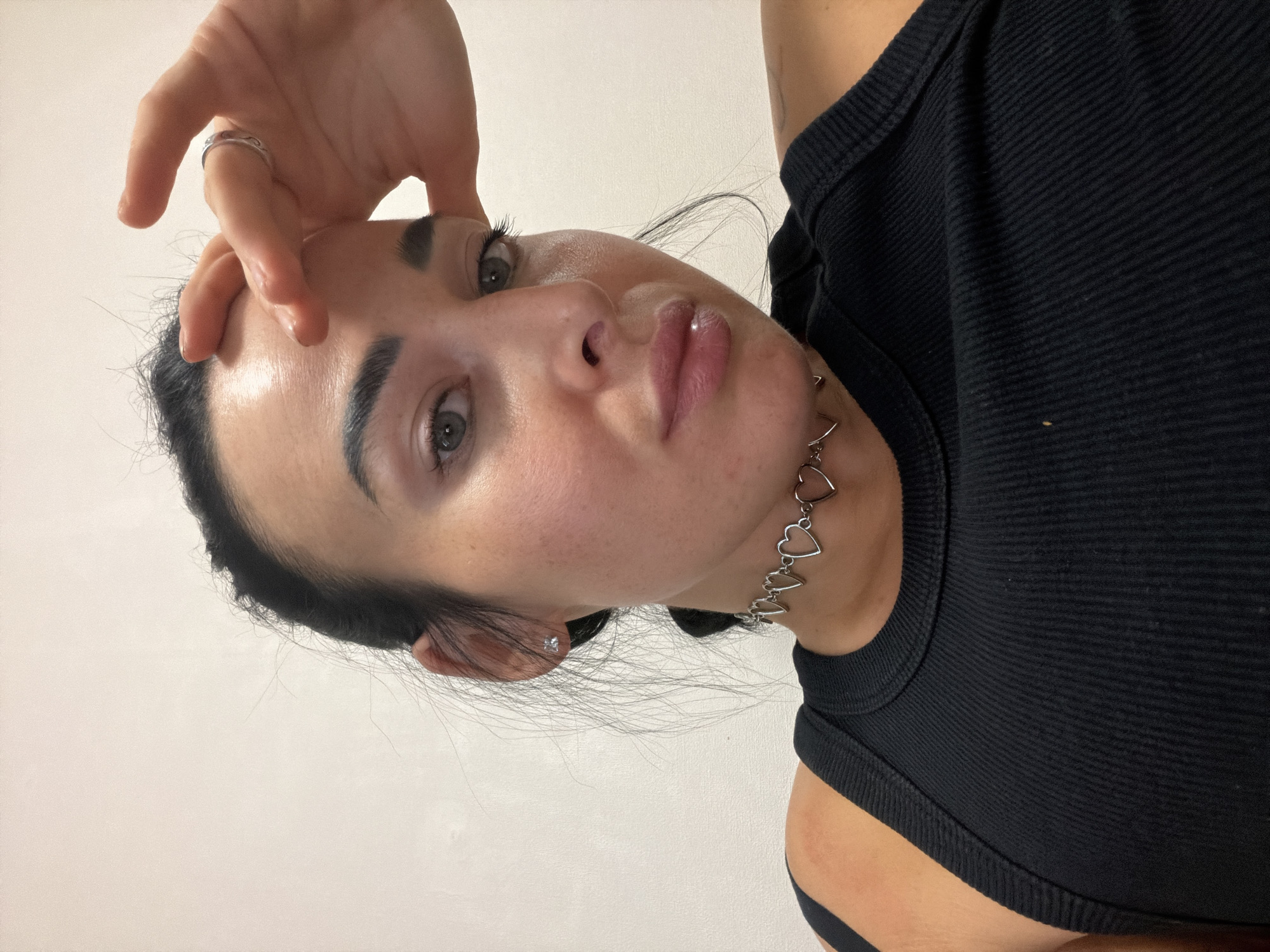 A headshot of a woman is presented from a low angle, rotated to the left by approximately 90 degrees. The woman has fair skin, dark hair pulled back, and expressive eyebrows. She is wearing a black ribbed tank top and a silver necklace with heart-shaped pendants. Her hand is raised to her forehead, with a ring visible on one of her fingers. The background is a plain, off-white wall. The location is not discernible from the provided image.
