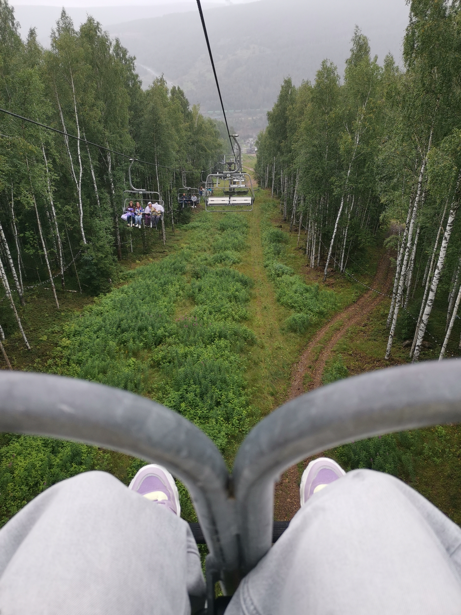 The image is taken from a cable car ascending a wooded hillside, likely near the city of Gubakha, Russia, known for its ski resorts and natural attractions. The perspective is from the viewpoint of a person sitting in the cable car, with their legs and feet visible in the foreground, wearing light grey pants and purple and white sneakers. The cable car itself, with its metal bar, is also in the foreground.
In the middle ground, the cable car track extends into a dense forest of birch trees with white bark and green foliage. Several other cable car cabins are visible on the track ahead, some carrying passengers. In one cabin, a group of four people is seated. Below the cable car track, a verdant, overgrown clearing is visible, with patches of grass and various green shrubs. A dirt path winds through the vegetation.
In the background, the forest continues up the hillside, fading into a hazy, overcast sky. The overall scene suggests an outdoor recreational activity, possibly a scenic ride during the warmer months. The overcast weather contributes to a slightly muted color palette.