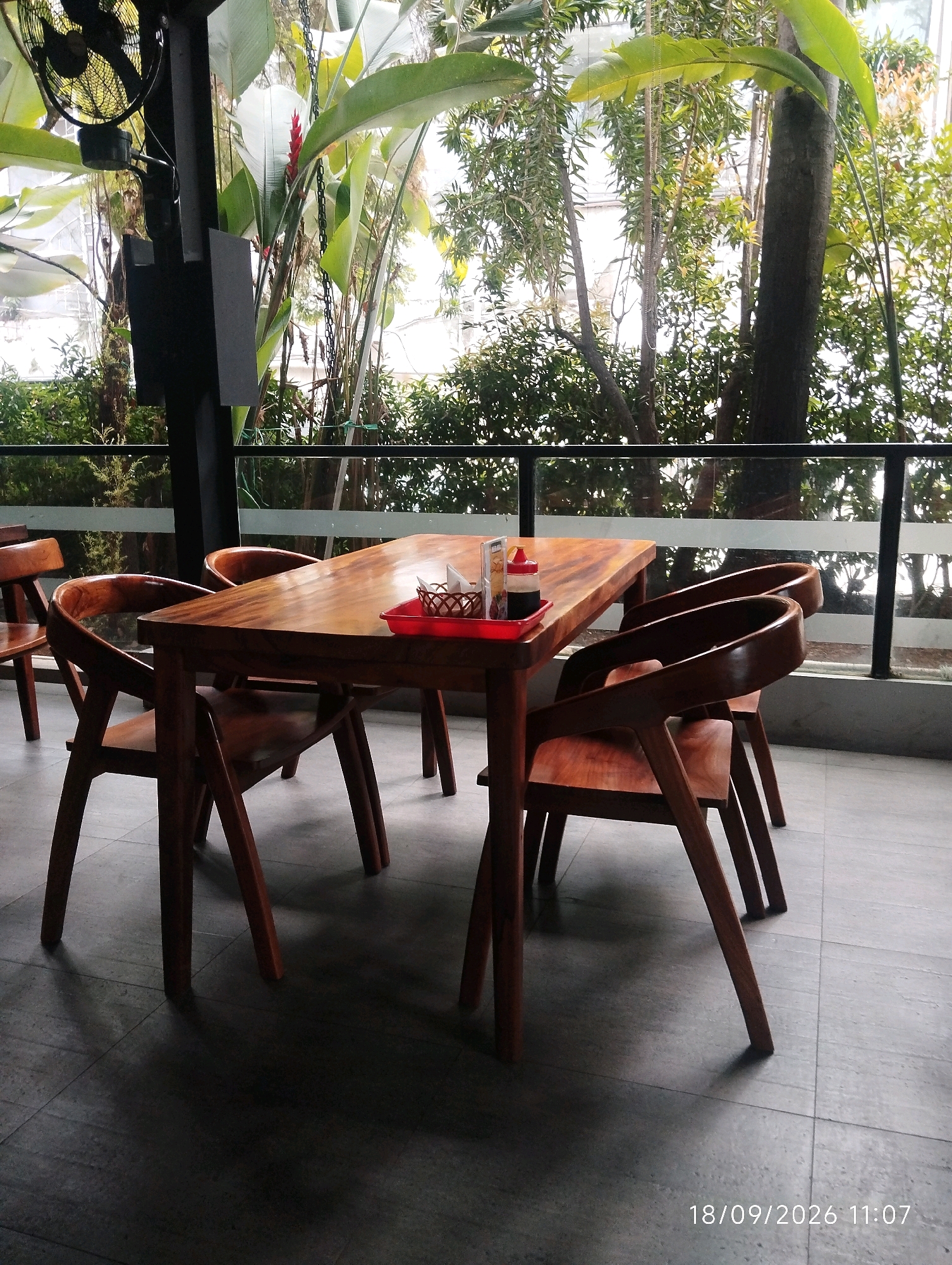The image depicts an outdoor seating area, likely a cafe or restaurant patio, with a wooden table and chairs set against a backdrop of lush greenery. The table is set with a red tray containing a small woven basket and two condiment bottles, one red and one dark brown, topped with white caps. The chairs are made of polished wood with curved backs. Behind the table, a railing separates the seating area from the outdoors, beyond which trees and foliage are visible. A large black fan is mounted on a black pole to the left of the frame, and a glimpse of a red flower is seen among the plants. The flooring is a dark grey tile. The timestamp "18/09/2026 11:07" is visible in the lower right corner. The scene appears to be set in a tropical or subtropical environment, characteristic of a city like Bandung, Indonesia, known for its cool climate and abundant greenery. There are no people visible in the image.