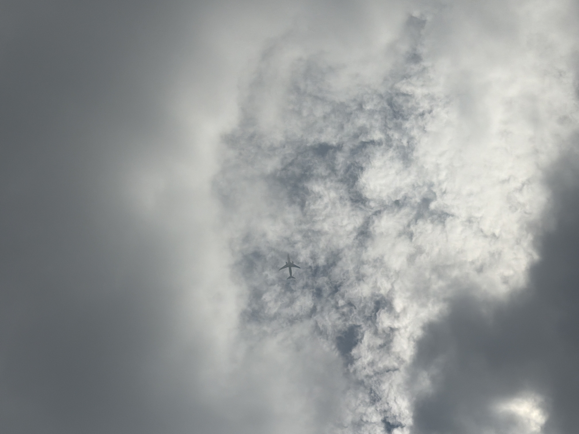 An airplane is visible flying through a cloudy sky above Panyu District, China. The image captures a vast expanse of overcast conditions, with layers of gray and white clouds dominating the frame. The airplane, a small dark silhouette against the lighter clouds, is positioned towards the upper middle section of the image. Its wings are extended horizontally, and its fuselage is elongated. The clouds are varied in texture and density, with some appearing wispy and translucent, while others are more substantial and shadowed. The overall lighting suggests a diffused daylight, typical of a cloudy day, with no direct sunlight piercing through. The scene is devoid of any other visible objects or people.