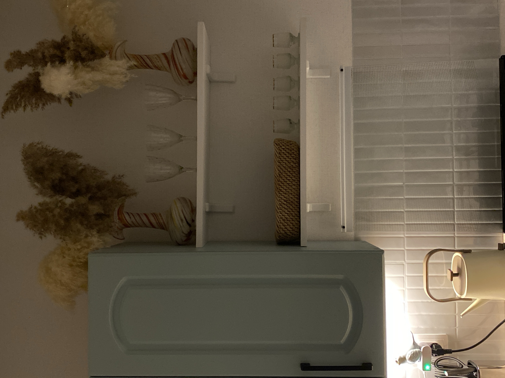 This is a close-up, eye-level, medium shot of a kitchen scene, possibly in Votkinsk, Russia. The foreground features a cream-colored kettle with a wooden handle and lid, positioned on a white tiled backsplash. To the left of the kettle, a small, white bird figurine is perched on a white electrical outlet adapter, which has a green light indicator. Next to the adapter, a silver utensil holder contains kitchen tools, including a pair of scissors. A black electrical cord connects to the adapter and extends out of frame.
Above the backsplash, two white shelves are mounted on a light-colored wall. The upper shelf holds two decorative vases with a striped pattern of red, white, yellow, and brown, each filled with dried pampas grass. Between the vases, four clear wine glasses are arranged. The lower shelf contains a woven basket and five small clear glasses, each with a gold rim.
To the left of the shelves, a portion of a light blue cabinet is visible, featuring a curved, recessed panel and a black handle. The lighting in the scene is warm and ambient, casting soft shadows. The overall impression is a neatly arranged kitchen counter area with decorative elements.