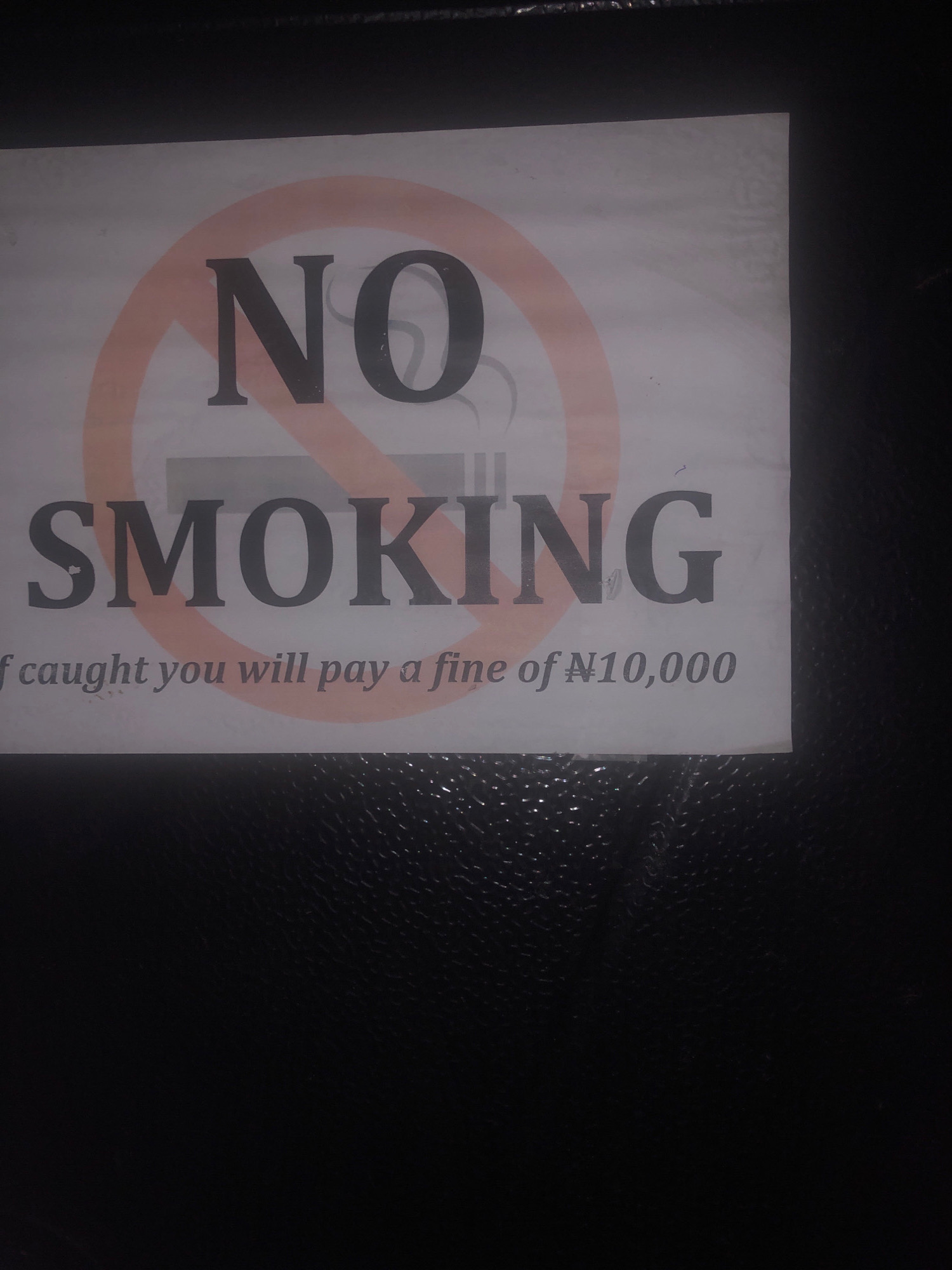 A "NO SMOKING" sign is posted on a dark, textured surface, likely a wall or door, in Etete, Nigeria. The sign is white with a red circle and diagonal line symbol over a silhouette of a cigarette. The words "NO SMOKING" are printed in large, dark capital letters below the symbol. Underneath, smaller text reads "f caught you will pay a fine of #10,000". The lighting suggests an indoor environment, possibly a public building or establishment.
