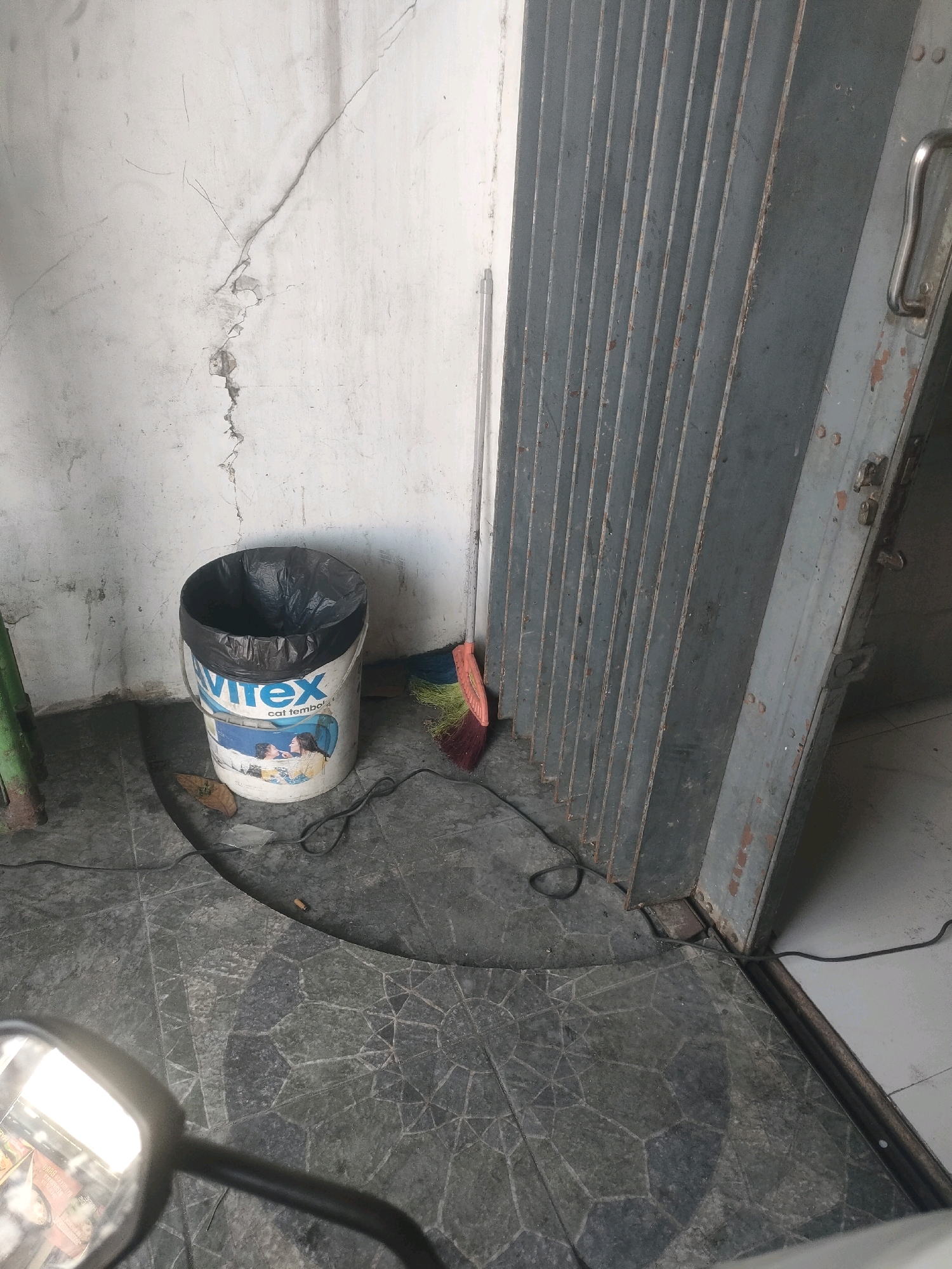 A paint bucket, a broom, and a motorcycle mirror are visible in this image, which appears to be taken in a low-lit, urban outdoor area. The background consists of a rough, cracked concrete wall and a corrugated metal roll-up door. The floor is paved with dark, irregularly shaped stones. A black plastic trash bag is inside the paint bucket, which has the brand name "Virex" and text "cat tembo" visible on its side. The broom has an orange handle and multi-colored bristles. The motorcycle mirror reflects the scene, showing a portion of what appears to be a store or building interior with lights and signage. The overall impression is one of a utilitarian, possibly neglected, outdoor space in Surabaya, Indonesia.