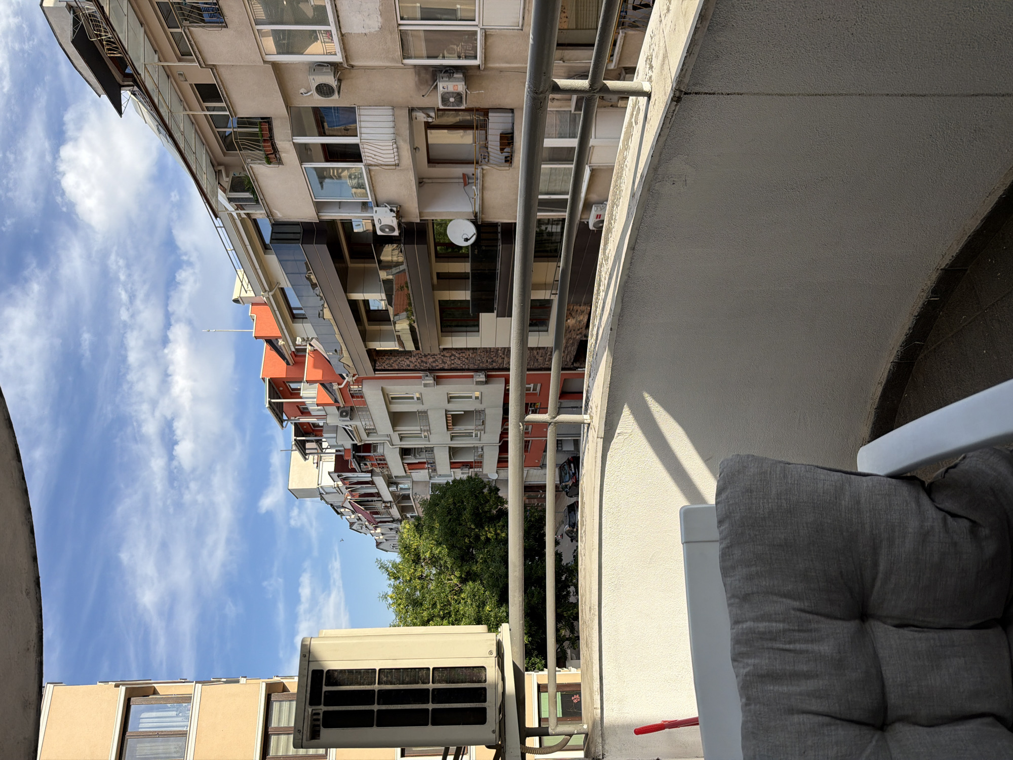 This is a photograph taken from a balcony in Plovdiv, Bulgaria, looking out onto a street lined with multi-story buildings. The sky is blue with scattered white clouds. On the balcony in the foreground, there is a cushioned chair and a red object, possibly a broom handle, leaning against the wall. A white air conditioning unit is mounted on the exterior wall of a building to the left. The buildings across the street are of varying architectural styles and colors, featuring balconies with railings and numerous windows. Several air conditioning units are visible on the building facades. A few cars are parked on the street below. Green trees are visible between the buildings. The lighting suggests it is daytime, likely late afternoon due to the shadows cast.