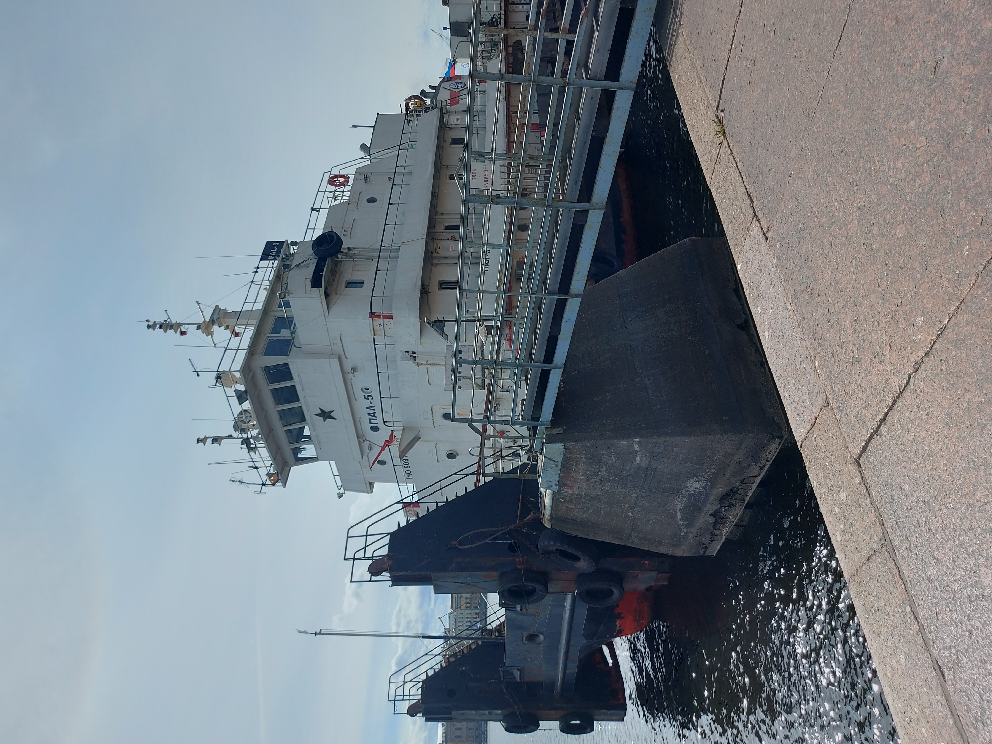 The image shows a ship docked along a granite embankment. The ship is a white vessel with multiple decks and visible navigational equipment. Text on the ship reads "OAI-5," and a star is painted near the windows of the bridge. A secondary barge-like structure is attached to the side of the ship, with tires acting as bumpers. The granite embankment is adjacent to dark water, reflecting the sky and the ship. A crane is visible in the background, as well as what appear to be buildings of Saint Petersburg. The sky is overcast.
