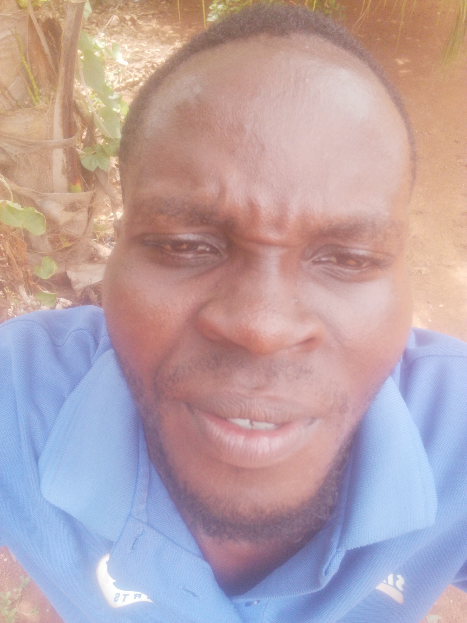 A close-up, high-angle portrait of a dark-skinned man wearing a blue collared shirt. His face is centered, with a slight frown, looking down at the camera. A short beard is visible on his face. Behind him, out of focus, is foliage, possibly a tree and green plants. The location appears to be outdoors. The city and state are Unknown, Unknown.