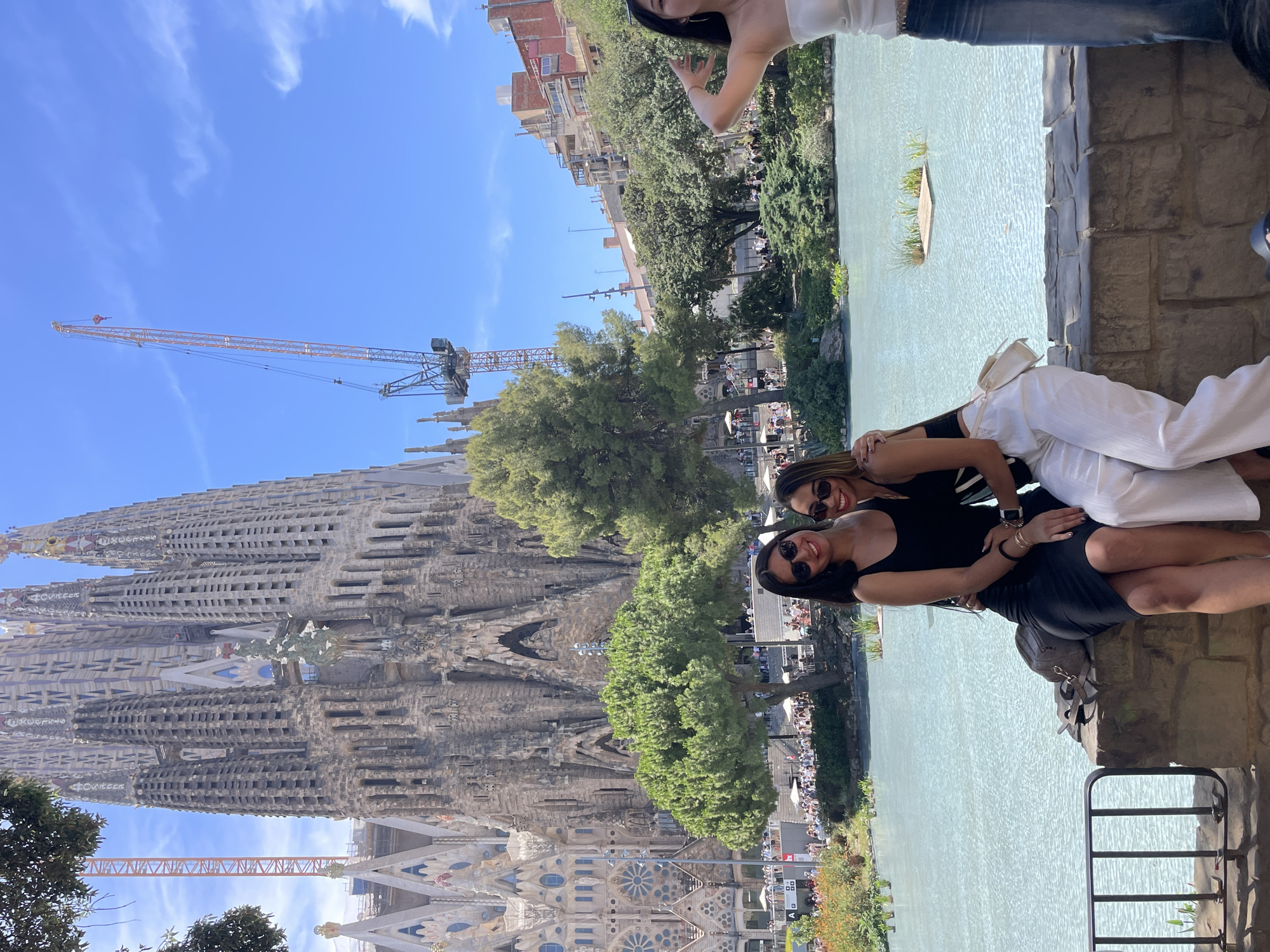 The image depicts two individuals posing for a photo in front of a large, intricately designed building, likely the Sagrada Familia in Barcelona, Spain, given the architectural style. The building is characterized by its numerous spires and detailed facades, with construction cranes visible in the background, indicating ongoing work. The two subjects are a woman in a black tank top and a woman in a white outfit, both wearing sunglasses and smiling at the camera. They are seated on a stone ledge overlooking a body of water that appears to be a large fountain or pool.
The scene suggests a sunny day with a clear blue sky and scattered white clouds. The time of day is likely midday or afternoon, judging by the bright sunlight and shadows. The individuals appear relaxed and happy, enjoying their time outdoors. The presence of other people in the background, along with trees and a cityscape, further places the scene in a public, tourist-oriented location. The overall atmosphere is one of leisure and sightseeing in a popular urban environment. No visible text is present in the image.