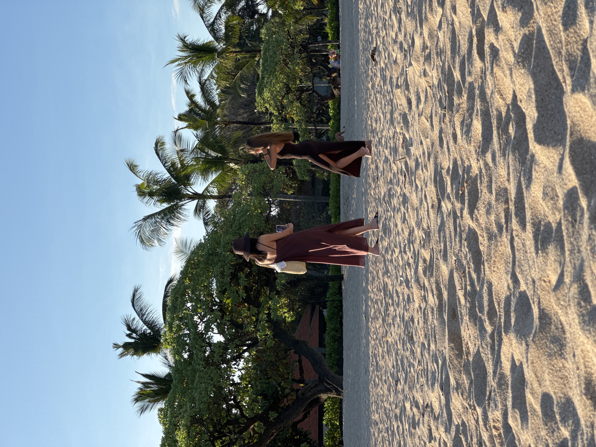 Two women stand on a sandy beach in Kuta Selatan, Indonesia, under a clear blue sky. The beach is lined with palm trees and other tropical vegetation.
The woman closest to the camera holds a phone, possibly taking a photo. Both women are wearing long dresses and wide-brimmed hats. The woman holding the phone has a small straw bag hanging from her shoulder. The sand is light brown.
