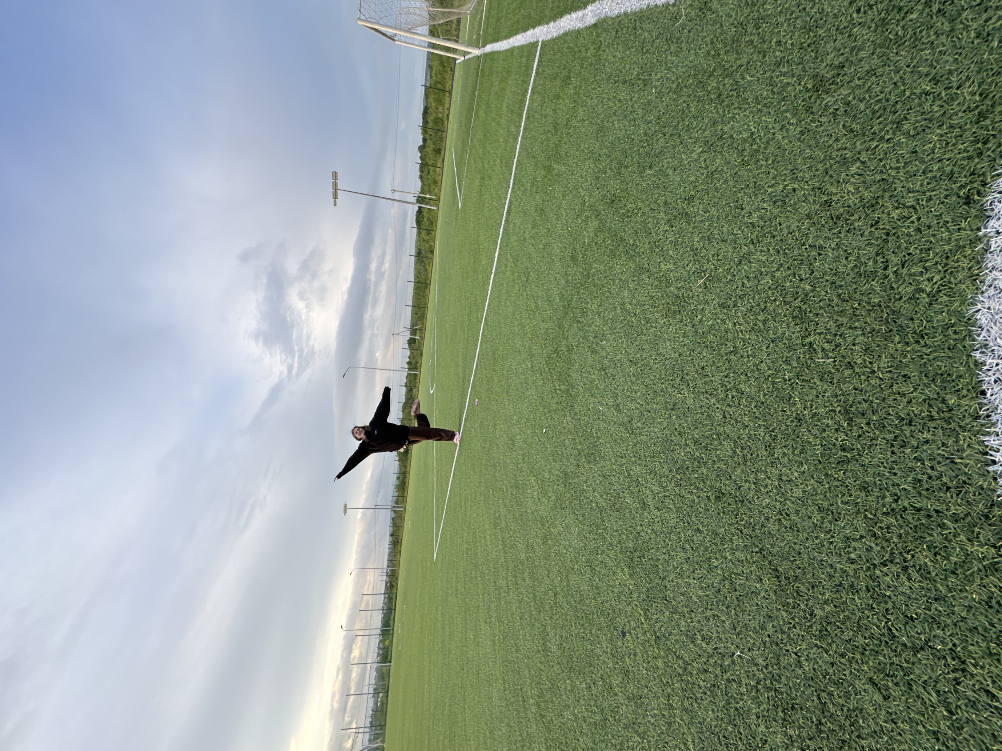 The image captures an artificial turf football field in the city of Ферма, Russia, under an overcast sky. A single individual, appearing to be a young female, stands in the mid-ground, balancing on her right leg with her left leg bent and elevated behind her. Her arms are extended laterally and angled slightly upward. She is dressed in a dark long-sleeved top, dark trousers, and light-colored footwear. A smile is discernible on her face. The field surface is green artificial turf marked with white lines, and the upper right corner of a white-netted goal post is visible on the right side of the frame. In the background, several tall light poles with floodlights are present, alongside a perimeter fence. Beyond the fence, a landscape of green vegetation and distant hills extends under the cloudy conditions.