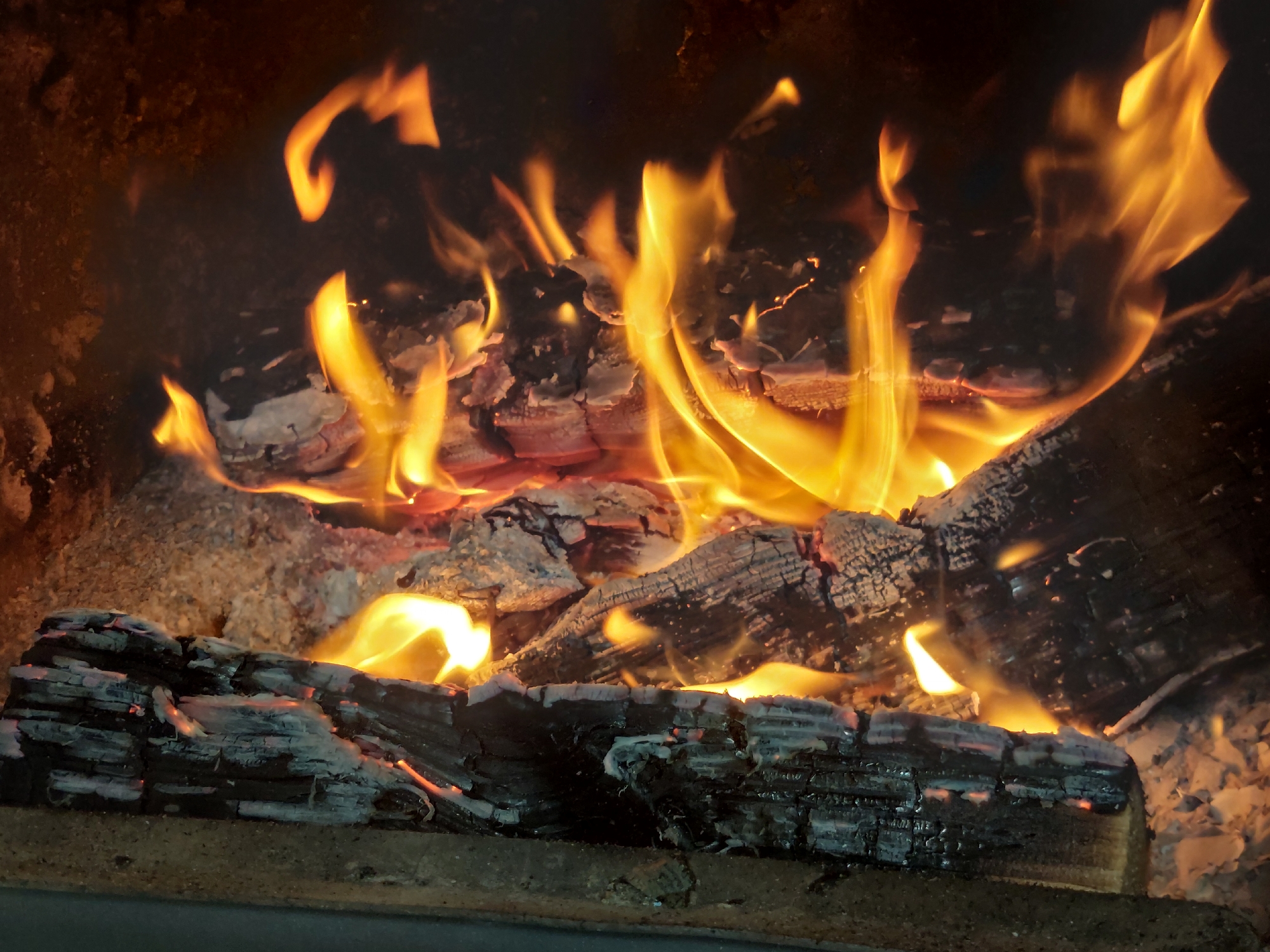 A close-up shot of a fire burning inside a fireplace. The logs are charred and surrounded by embers. Flames lick upwards, illuminating the dark interior of the fireplace. There is no indication of the location or people.