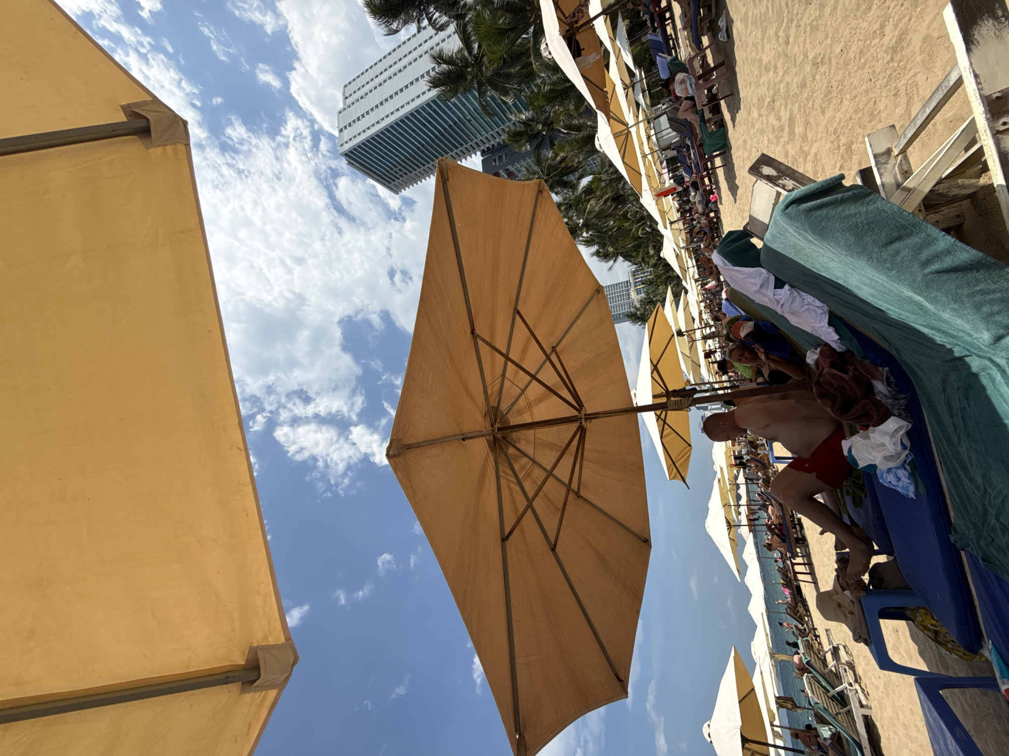 The image captures a vibrant beach scene in Nha Trang, Vietnam, under a bright, partly cloudy sky. In the foreground, a large, tan beach umbrella provides shade, its structure visible against the blue sky. Below the umbrella, a man in red swim trunks reclines on a blue beach chair, his legs extended. Adjacent to him, another beach chair is draped with green and white towels and a straw hat rests on a blue plastic side table.
The beach is lined with rows of similar beach chairs and umbrellas, some occupied by sunbathers. Palm trees dot the background, interspersed with several tall, modern buildings, including a prominent, light-colored skyscraper with dark vertical accents. The sand is golden and appears well-maintained. The overall atmosphere suggests a relaxing day at a popular tourist destination.