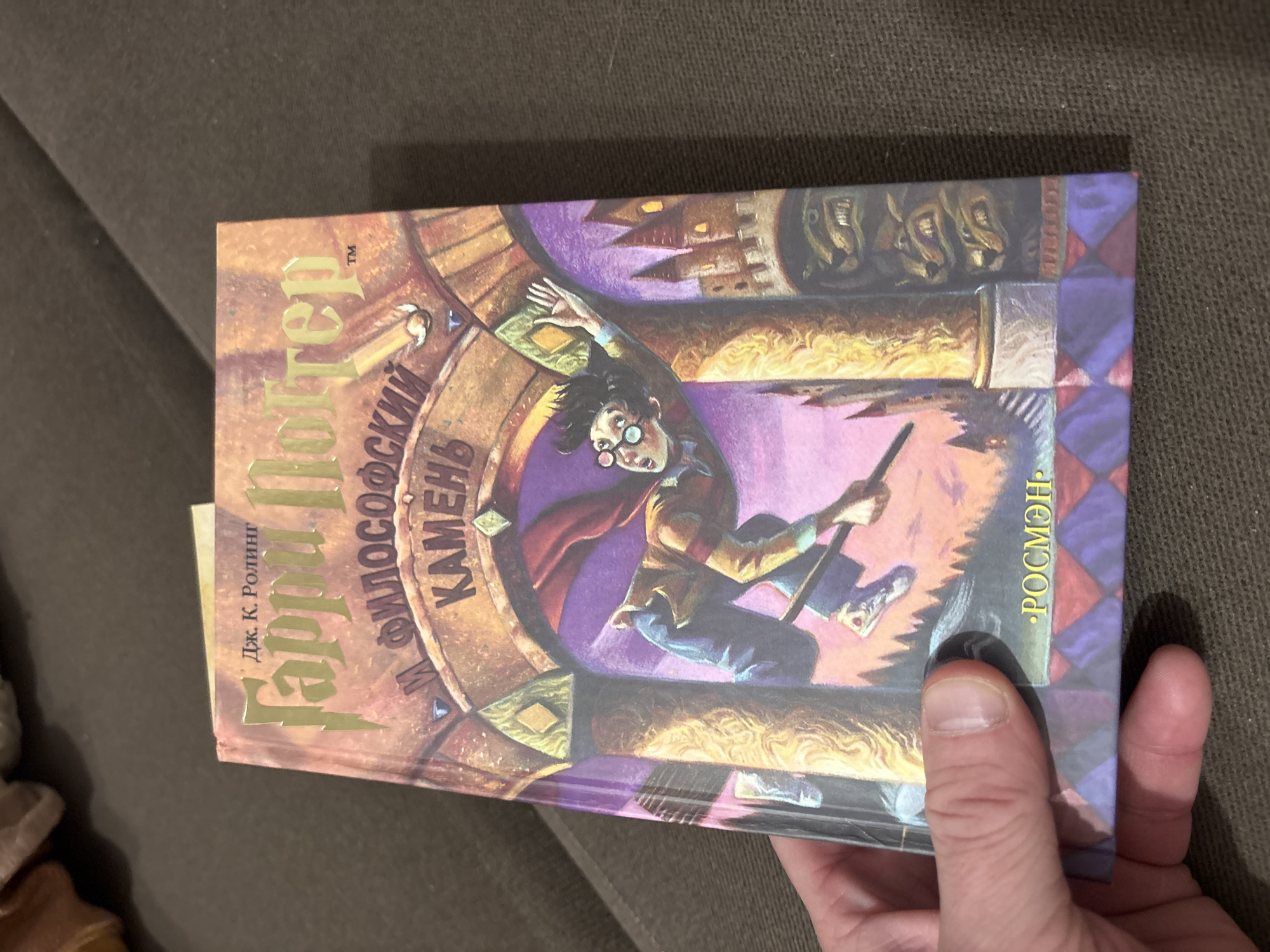 The image shows a copy of "Harry Potter and the Philosopher's Stone" (Гарри Поттер и философский камень) held in a hand. "РОСМЭН" is printed on the spine, indicating it's a Russian edition by Rosman publishing house. The book is not located in the city of Perm, Russia.