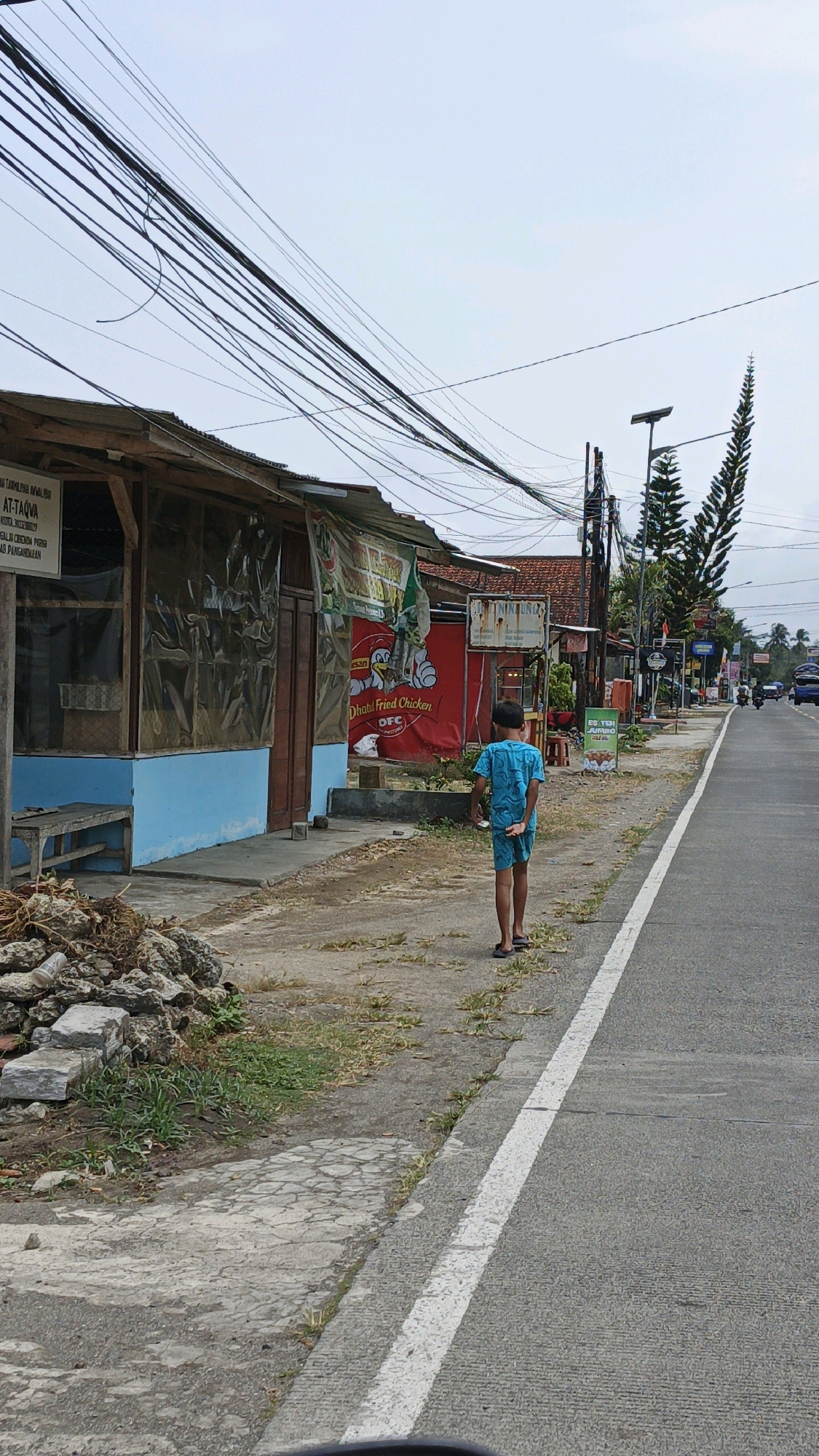 A young boy in a blue t-shirt and shorts walks away from the camera on the side of a paved road in Cibenda, Indonesia. He is walking on a dirt path that runs alongside the road, which has a white stripe painted down the center. To his left, there is a low-slung building with a corrugated metal roof and a blue painted lower section. This building has a wooden door and windows covered by plastic sheeting. A wooden bench sits in front of the building. Piles of rocks and debris are scattered along the edge of the path. Across the road, there are more buildings and shops, including a red banner advertising "Dhati Fried Chicken". The sky is overcast. Power lines stretch across the top of the frame.