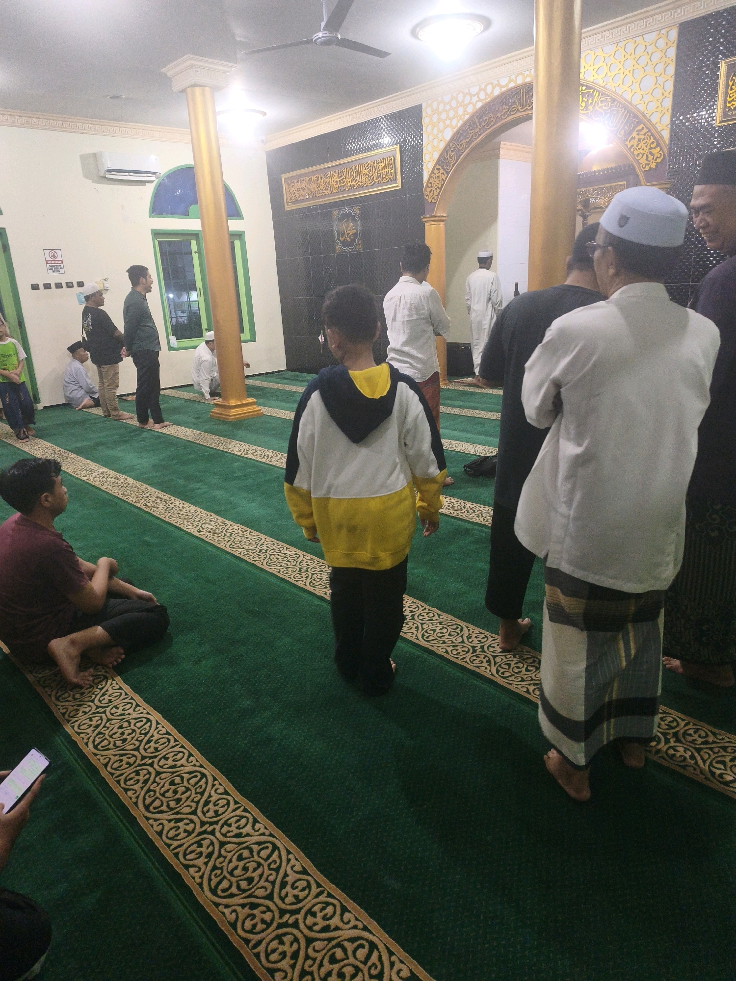 The image depicts the interior of a mosque in Samarinda, Indonesia. Several individuals are present, many of whom are men and boys. The floor is covered with a green carpet featuring ornate gold trim. In the foreground, a boy is seated on the floor, looking towards the right. Another boy, wearing a yellow and white hooded jacket, stands facing away from the camera. In the mid-ground, men are standing, some in conversation, and others appear to be observing. One man is wearing a white skullcap and a white shirt, while another is in a black shirt and pants. Further back, men are seen in prayer or preparation for prayer. The mosque's architecture includes arches, pillars, and decorative wall coverings with Arabic calligraphy. An air conditioning unit is visible on the wall. The lighting appears to be artificial, emanating from overhead fixtures. The overall atmosphere suggests a religious gathering or prayer session within the mosque.