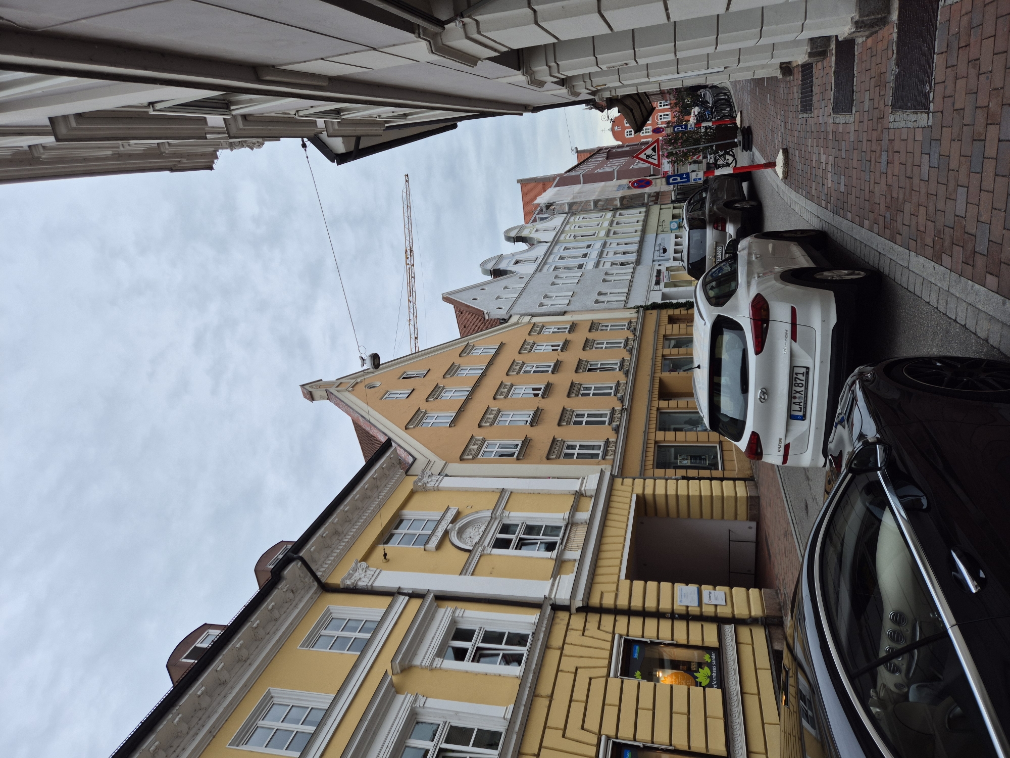 The image depicts a street scene in the city of Landshut, Germany. On the left side of the street, there are yellow buildings with traditional architecture, featuring white window frames and decorative elements. The buildings have multiple stories and a textured facade. An archway is visible between two of these buildings, leading into a darker interior.
On the right side of the street, the architecture is different, with lighter-colored buildings featuring a more classical style, characterized by columns and elaborate cornices. The street is paved with red brick.
Several cars are parked along the street. In the foreground, a black luxury car is partially visible on the left, with its interior showing. Further down the street, a white SUV is parked, displaying a German license plate. Another car, a silver SUV, is parked behind it.
Various street signs are present, including parking signs and a construction sign. A bicycle rack with bicycles is also visible. In the background, a construction crane can be seen extending over the rooftops. The sky is overcast, suggesting a cloudy day. The overall impression is of a historic European street in an urban setting, with elements of both residential and commercial buildings.