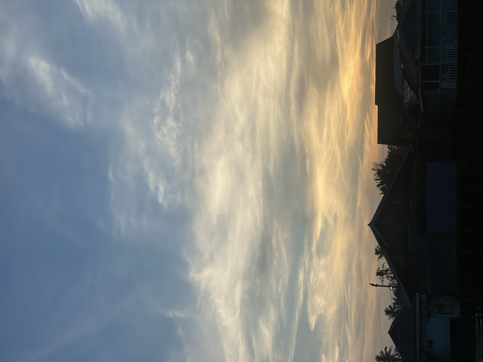 The image depicts a collection of silhouetted houses and palm trees against a vast, cloudy sky. The sky transitions from a soft blue at the top to warm orange and yellow hues near the horizon, suggesting either a sunrise or sunset. The houses, dark and indistinct, appear to be built on stilts, a common architectural feature in flood-prone areas. The overall scene evokes a peaceful, possibly rural, atmosphere. This scene is located in the city of Banjarmasin, Indonesia, a region known for its extensive river systems and stilt housing.