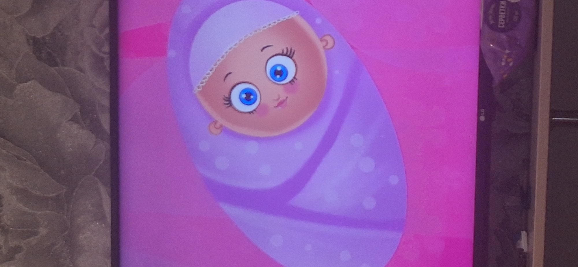 The image shows a television screen displaying a cartoon of a baby. The baby has large blue eyes with long eyelashes and a small pink mouth. It is wrapped in a purple blanket with white polka dots and wearing a white bonnet. The background is a vibrant pink. At the bottom of the screen, the LG logo is visible, along with a package of what appears to be wet wipes with Russian text that translates to "SERBETKI" (wipes). The television is placed on a shelf above a wooden surface. The image does not contain any information or visual cues that would allow for its location to be identified in Bila Tserkva, Ukraine.