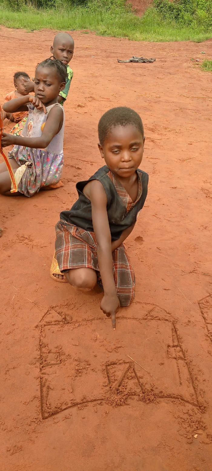 In the city of Ngalula, Democratic Republic of the Congo, a group of children is observed outdoors on a reddish-brown dirt surface. In the foreground, a young child, appearing approximately 5-7 years old, with short dark hair and wearing a dark sleeveless top over a plaid skirt, kneels and points with their right index finger at drawings etched into the dirt. These drawings include the outline of a house and what appears to be the capital letter 'A'. To the left and slightly behind, another child with braided hair, wearing a light-colored patterned dress, sits on the ground, holding a red and yellow strap-like object. Further back and slightly to the right, a third child with a shaved head and a green patterned shirt looks directly forward. A baby or very young child is partially visible to the far left, behind the child in the patterned dress. The background consists of more reddish-brown dirt, with green foliage and bushes visible in the distance, suggesting an open, unpaved area. Scattered detritus is present on the ground in the mid-background.