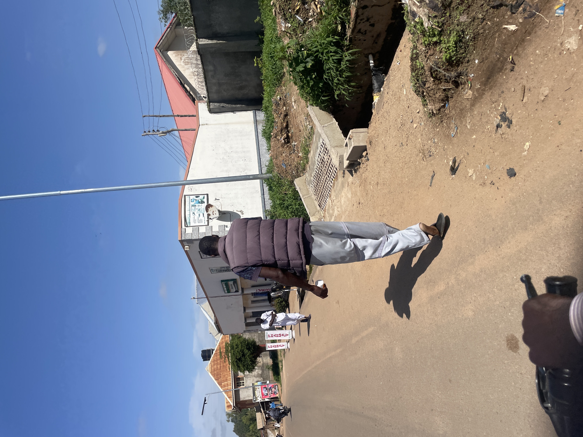 The image shows a street scene in Jos, Nigeria. A man is walking away from the camera on a dirt road. He is wearing a dark vest over a patterned shirt and light-colored pants. He has a white object in his right hand. In the background, there are buildings with red roofs, a street light, and power lines. To the right of the road, there is a drainage ditch filled with water and debris. The sky is blue with some scattered clouds. The scene appears to be a typical street in an urban or semi-urban area.