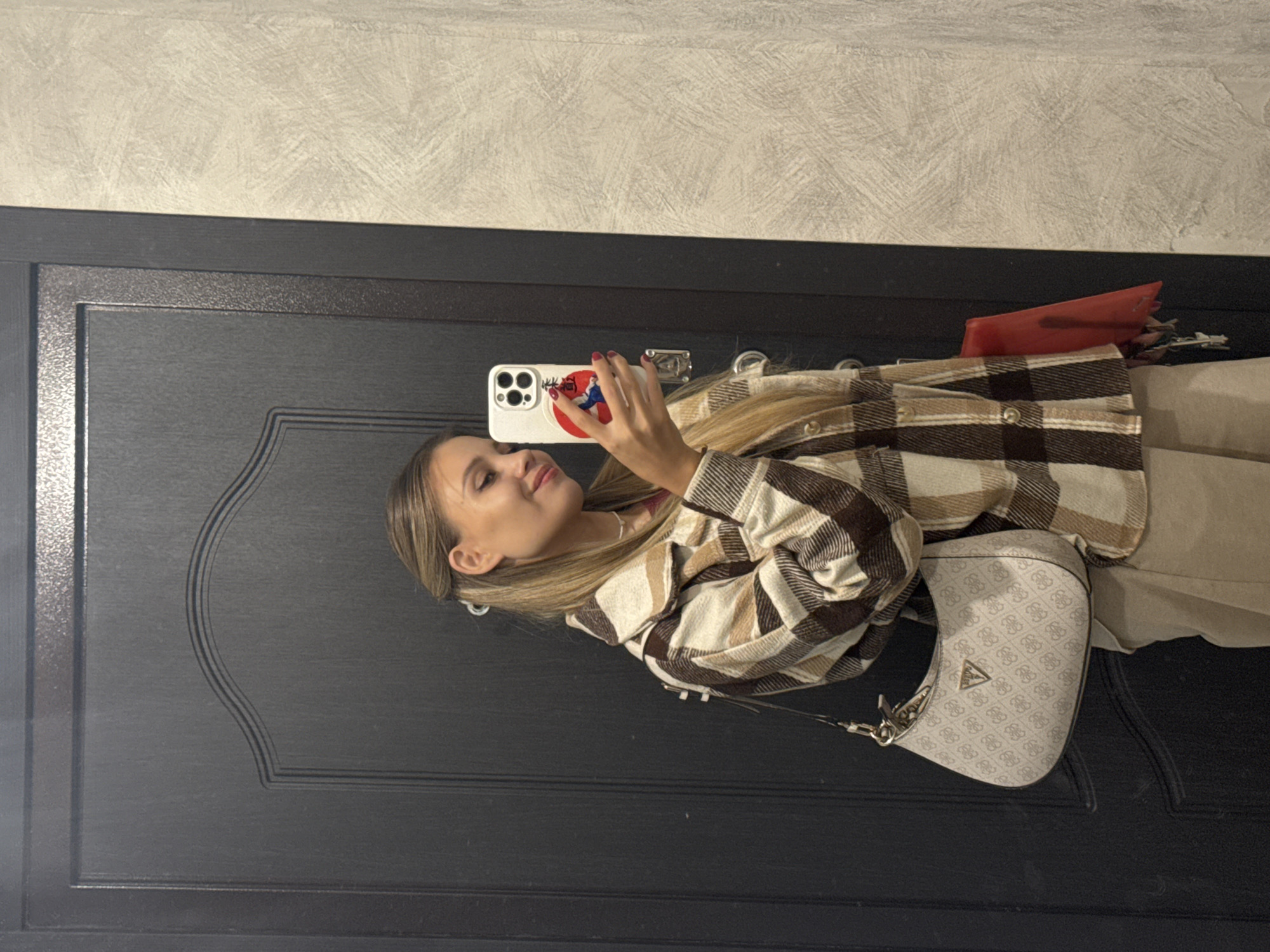 A young woman with blonde hair styled back with a clip is taking a selfie in a doorway. She is wearing a brown and white plaid shirt jacket over a dark red shirt, and beige pants. She is carrying a beige Guess handbag with the brand's logo pattern. In her right hand, she holds a white smartphone with a decorative case. A red bag is tucked under her left arm, and keys are visible hanging from it. The background consists of a dark wooden door and a textured grey wall. The scene appears to be indoors, possibly in an apartment or building hallway in the city of Chaikovsky, Russia.