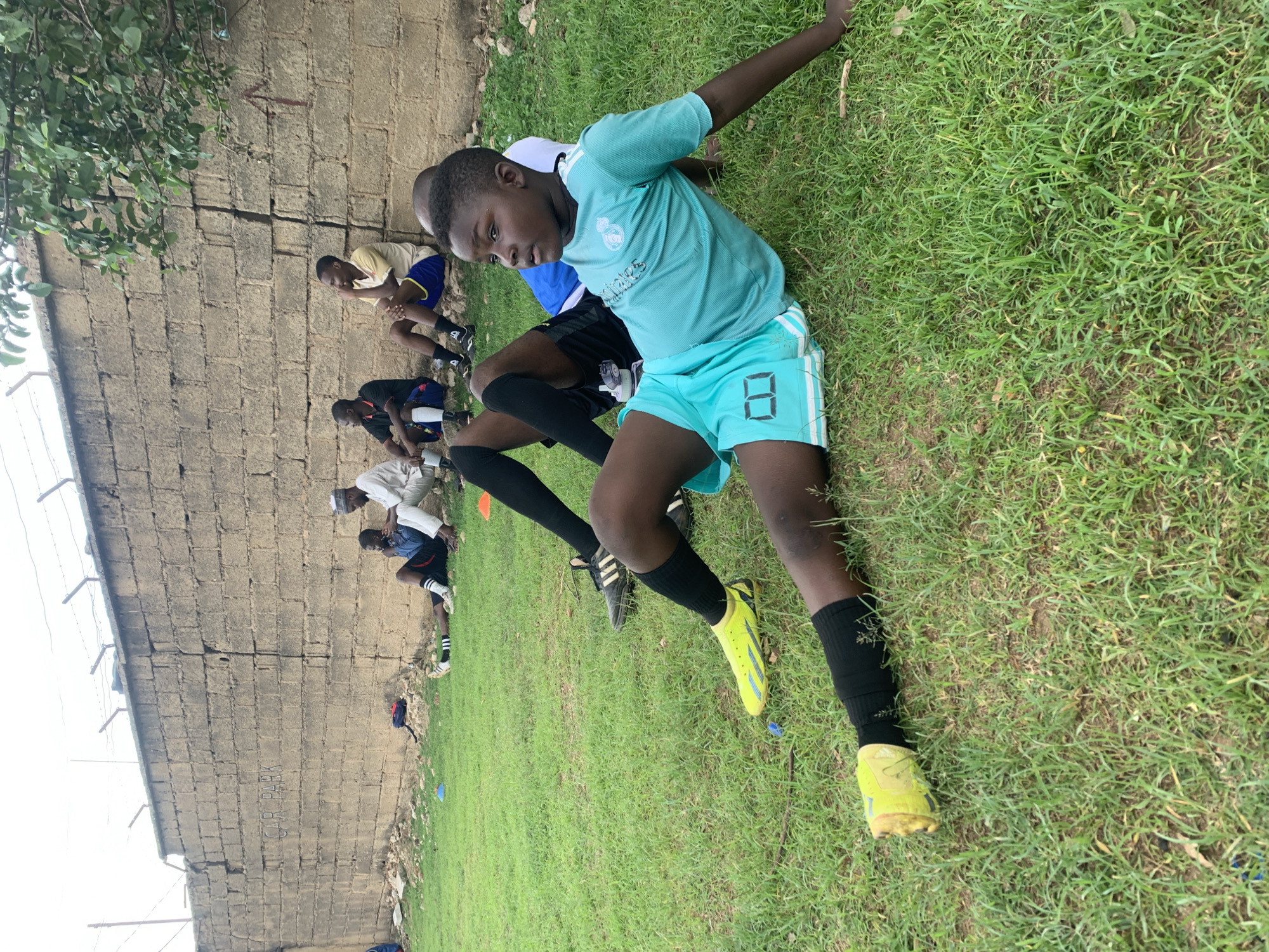 A group of young men are engaged in a soccer practice in Jos, Nigeria. They are positioned on a grassy field adjacent to a tall, weathered concrete block wall. The young man in the foreground wears a light blue soccer jersey and shorts with the number 8, black socks, and yellow cleats. Several other individuals are performing seated stretches along the wall. Overhanging greenery from a tree is visible in the upper left corner.