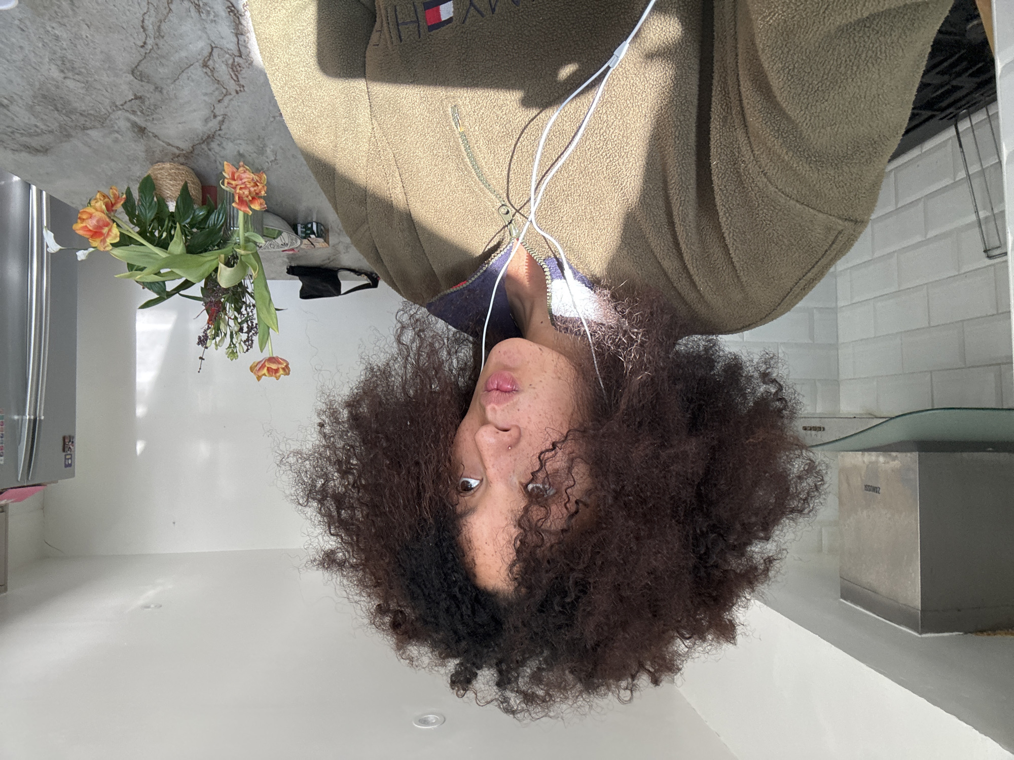 This media file presents an upside-down, close-up portrait of an individual in what appears to be an indoor setting in London, United Kingdom. The person has a youthful appearance, characterized by dark, voluminous curly hair and fair skin adorned with numerous freckles across their face. They are looking directly at the camera with light-colored eyes and a neutral, slightly contemplative expression, with their upper lip visibly pierced.
The individual is wearing a khaki-green fleece jacket featuring a quarter-zip design. A dark blue collar is visible beneath the fleece. On the upper chest of the jacket, the "TOMMY HILFIGER" logo is prominently displayed. White wired earphones are plugged into their ears, with the wires extending across their chest.
The background suggests a well-lit modern kitchen or dining area. To the viewer's left (or the subject's right), a vibrant floral arrangement is placed on a light grey, marbled countertop. The bouquet consists of lush orange and peach tulips, complemented by green foliage and smaller purple and white flowers, all held in a clear glass vase. Next to the vase, a small, woven, light-brown decorative pot or basket is visible. Further to the left, a sliver of a stainless steel appliance, possibly a refrigerator or dishwasher, can be seen. On the opposite side of the frame, a wall finished with white subway tiles extends upwards, and beneath it, a dark grey, block-like object is present, bearing the text "ZANUSSI". The scene is illuminated by bright natural light, casting distinct shadows on the jacket and the countertop, indicating it is daytime.