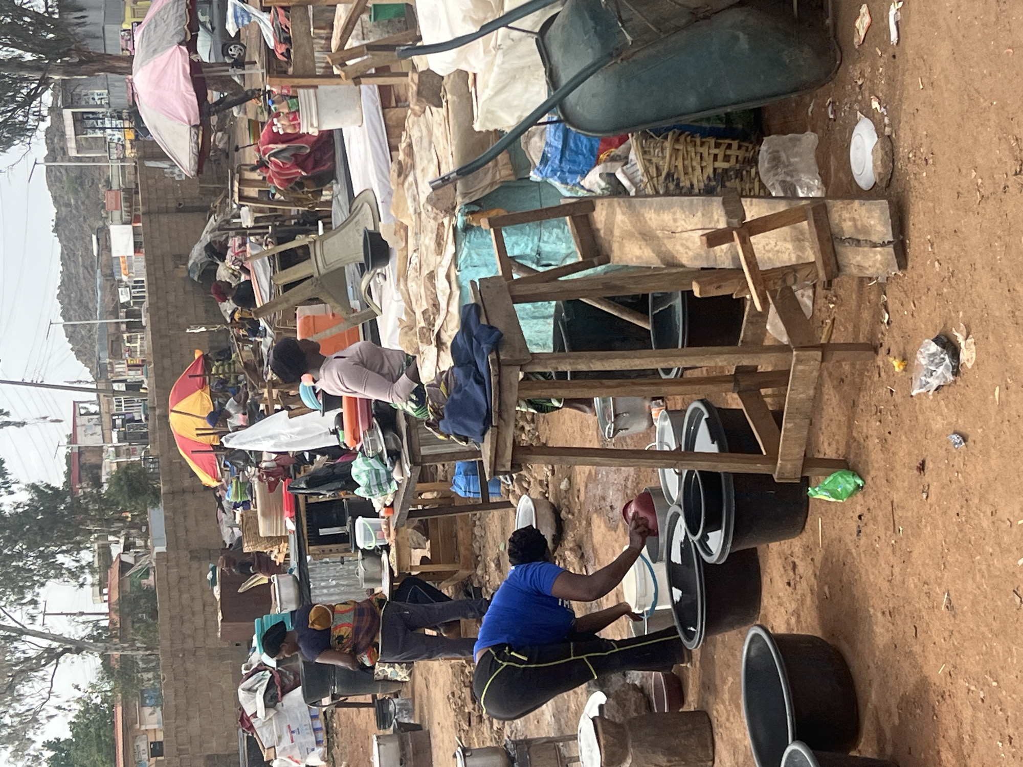 The scene depicts an outdoor setting in Gura Top, Nigeria, with a chaotic arrangement of market stalls and activity. Several people are present, including a woman in a blue shirt transferring liquid from a red container into one of the many black buckets lining the ground. Other individuals are seen nearby, seemingly engaged in similar activities amidst the cluttered environment. The area is densely packed with goods and makeshift structures, including wooden scaffolding and tarpaulins. The backdrop shows a partial view of buildings and landscape, suggesting a busy urban area.