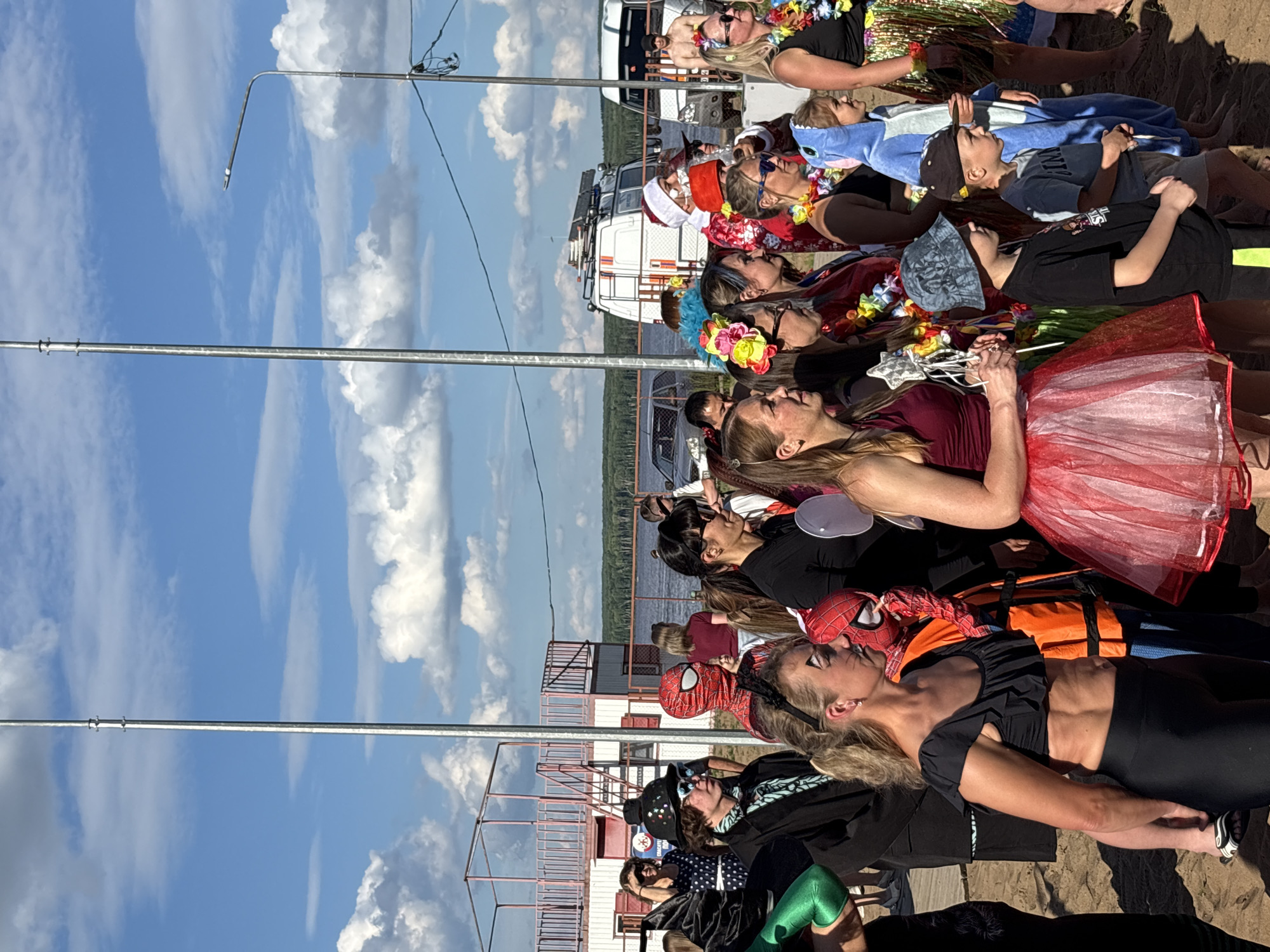 The image shows a group of people gathered on a sandy area, likely a beach or riverbank, with a body of water and trees in the background. The sky is blue with fluffy white clouds, suggesting a pleasant day. The people are dressed in a variety of outfits, many of them costumes, including a person in a Spider-Man suit, someone in a shark costume, and others wearing leis and festive accessories. Some individuals appear to be children, while others are adults. The scene suggests a casual outdoor gathering or event, possibly a festival or a themed party. The orientation of the image is vertical, and it seems to be taken during the daytime due to the bright lighting and clear skies. The location is identified as Chaikovsky, Russia. There are metal structures resembling bleachers or a pier in the background, and a white vehicle that looks like a truck or van. The overall mood appears to be festive and relaxed, with people posing for photos or engaging in activities.