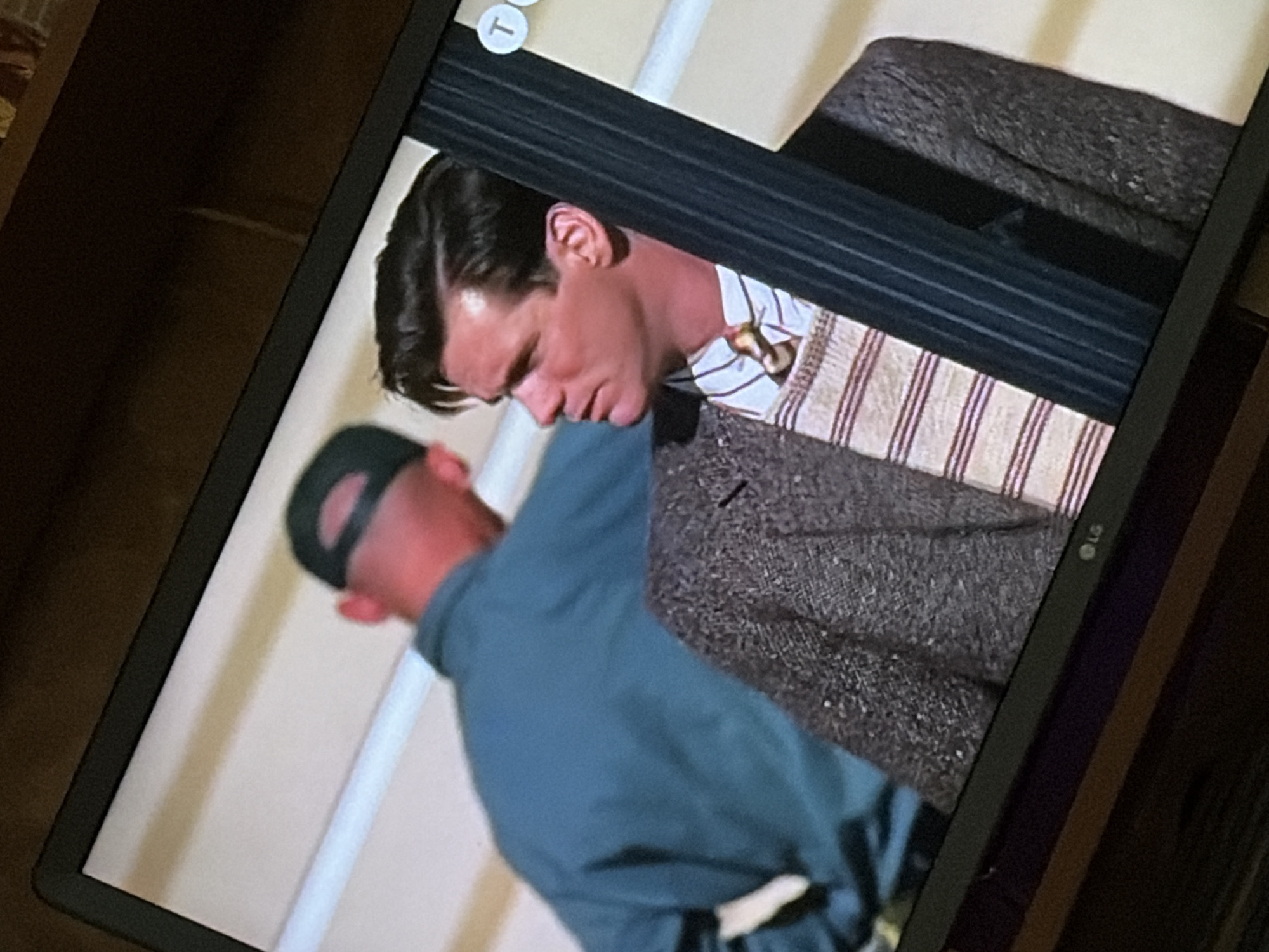 The image is a photograph taken from an angle, likely of a television screen displaying a scene. In the foreground, a man with dark, slicked-back hair and a light complexion is visible from the chest up, looking to his left. He is wearing a tweed jacket over a striped sweater and a tie. Behind him, another person wearing a dark baseball cap and a teal jacket is seen from the back. The background consists of a light-colored wall or structure with horizontal lines. The scene appears to be indoors, possibly on a set or a street with architectural elements. There is no indication of the location being Osolynka, Ukraine, or any other specific geographical place.