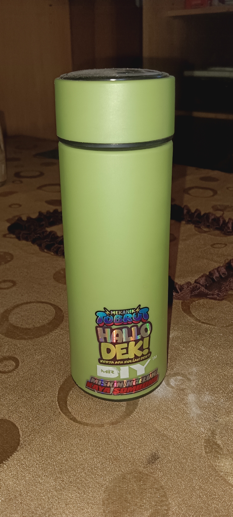 A light green, cylindrical thermos is positioned vertically on a patterned brown tablecloth. The thermos has a black rim at the top and a black band around its base. A sticker adorns the front of the thermos, featuring colorful, holographic text. The sticker's text reads "MEKANIK TOBRUT HALLO DEK! KERJA APA KULIAH DEK? DIY" and "MISKIN NGELUH KAYA SOMBONG". The background shows a blurred view of wooden shelves and a dark surface. The lighting is dim, and dust is visible on the top of the thermos.