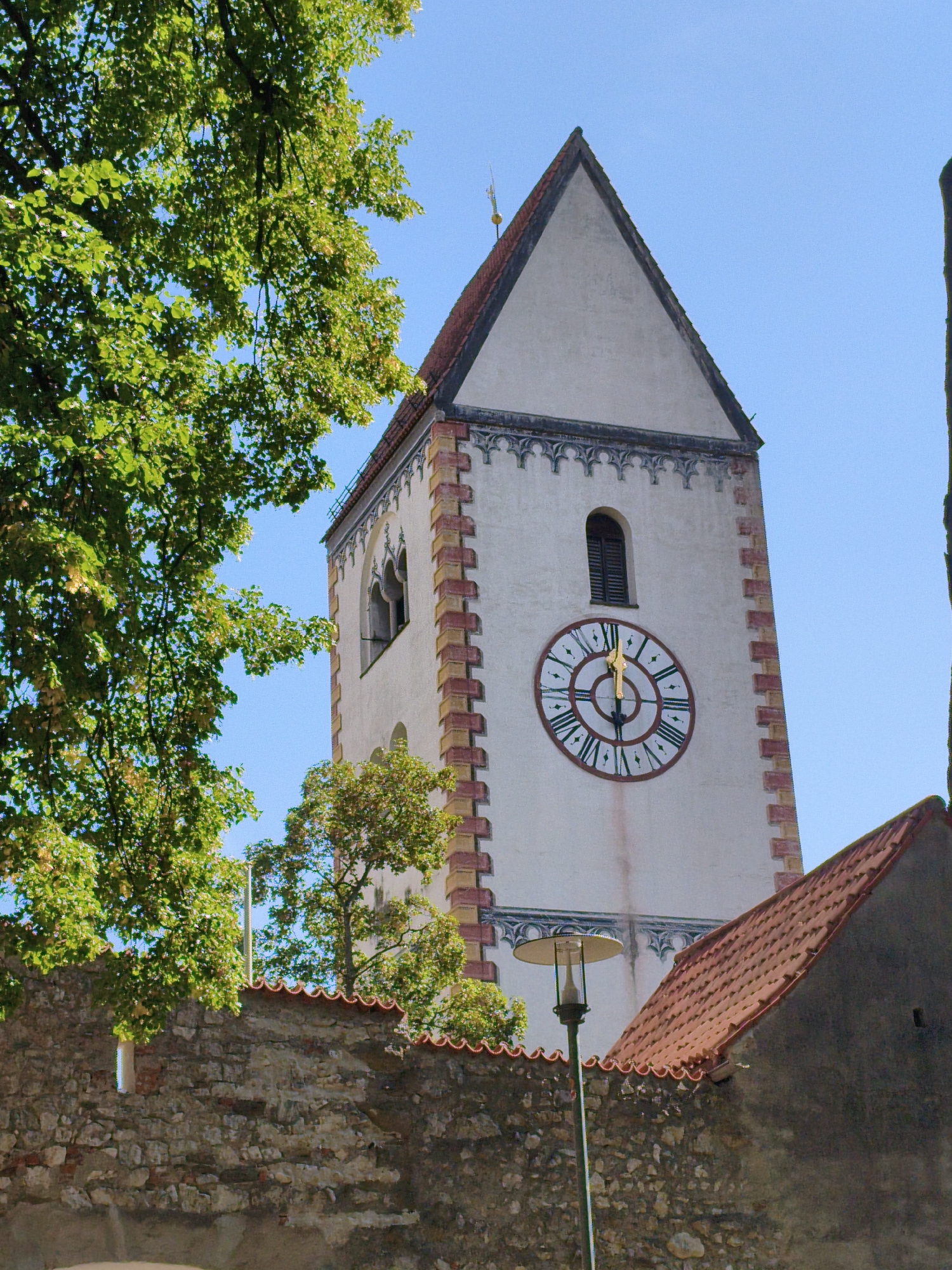 The image shows a detailed view of the tower of St. Mang's Abbey in Füssen, Germany, on a clear, sunny day. The tower, a prominent landmark, is tall and white with a pitched roof. It features a large, circular clock face with Roman numerals and ornate hands. Above the clock, a small, arched window is visible. Decorative friezes run along the upper sections of the tower. The tower is bordered by a textured, reddish-brown stone trim.
In the foreground, a weathered stone wall with visible bricks and mortar spans the lower part of the image. Lush green foliage from a large tree partially obscures the left side of the tower and the sky. To the right of the tower, a smaller building with a red-tiled roof is partially visible. A lamppost with a wide, circular shade stands near the stone wall, its base obscured. The sky is a vibrant blue and cloudless. The scene suggests a peaceful, historical setting within the city.