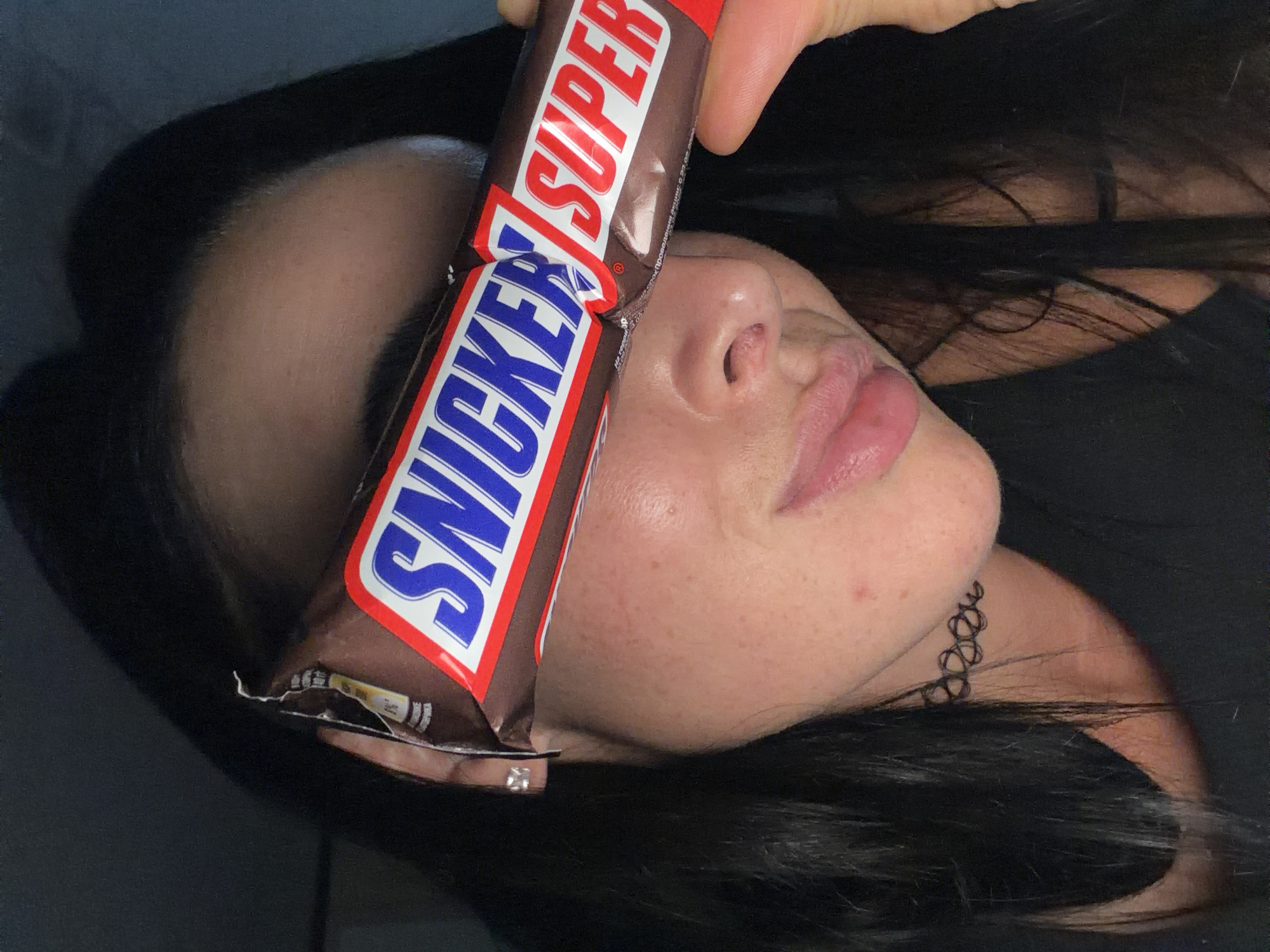 An image captures a person holding a Snickers Super chocolate bar horizontally across their face, effectively obscuring their eyes. The shot is angled, revealing the person's nose, mouth with full lips, and chin. They have dark hair, which is visible around the face and extends downwards, and fair skin with some freckles. A black choker-style necklace is visible on their neck/chin area.
The Snickers bar, a "Super" size, dominates the center of the frame. The iconic "SNICKERS" logo is prominent in blue letters on a white background, underlined by red, with "SUPER" in red letters above it. The wrapper appears slightly crinkled, suggesting it has been handled. The person's hand is visible at the top, holding the bar.
The overall lighting is soft and somewhat dim, possibly indicating an indoor setting or evening. The pose suggests a playful or candid moment, with the chocolate bar humorously replacing the eyes. There are no specific cues within the image itself to denote a location, but the context provided places the scene in Saint Petersburg, Russia.