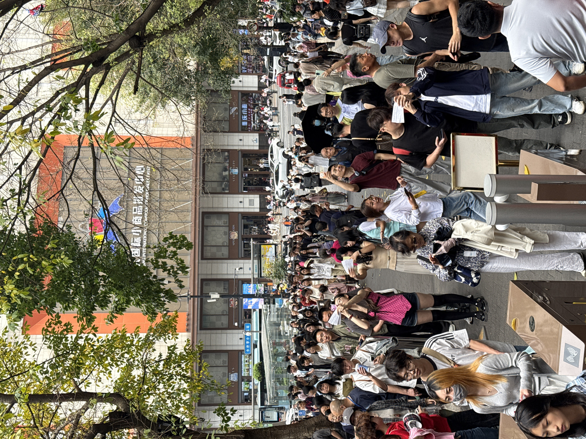 A daytime outdoor scene depicts a densely packed crowd of people lined up on a city street in Yuzhong District, China. The majority of the people are standing, but many are seated or lying down on the pavement while looking at their phones. The people are of various ages and wear casual clothing. Some are wearing face masks. The scene appears to be a queue, possibly for an event or attraction. Behind the crowd are buildings with storefronts, as well as overhead signs in Chinese and English. Trees with foliage are visible along the street.