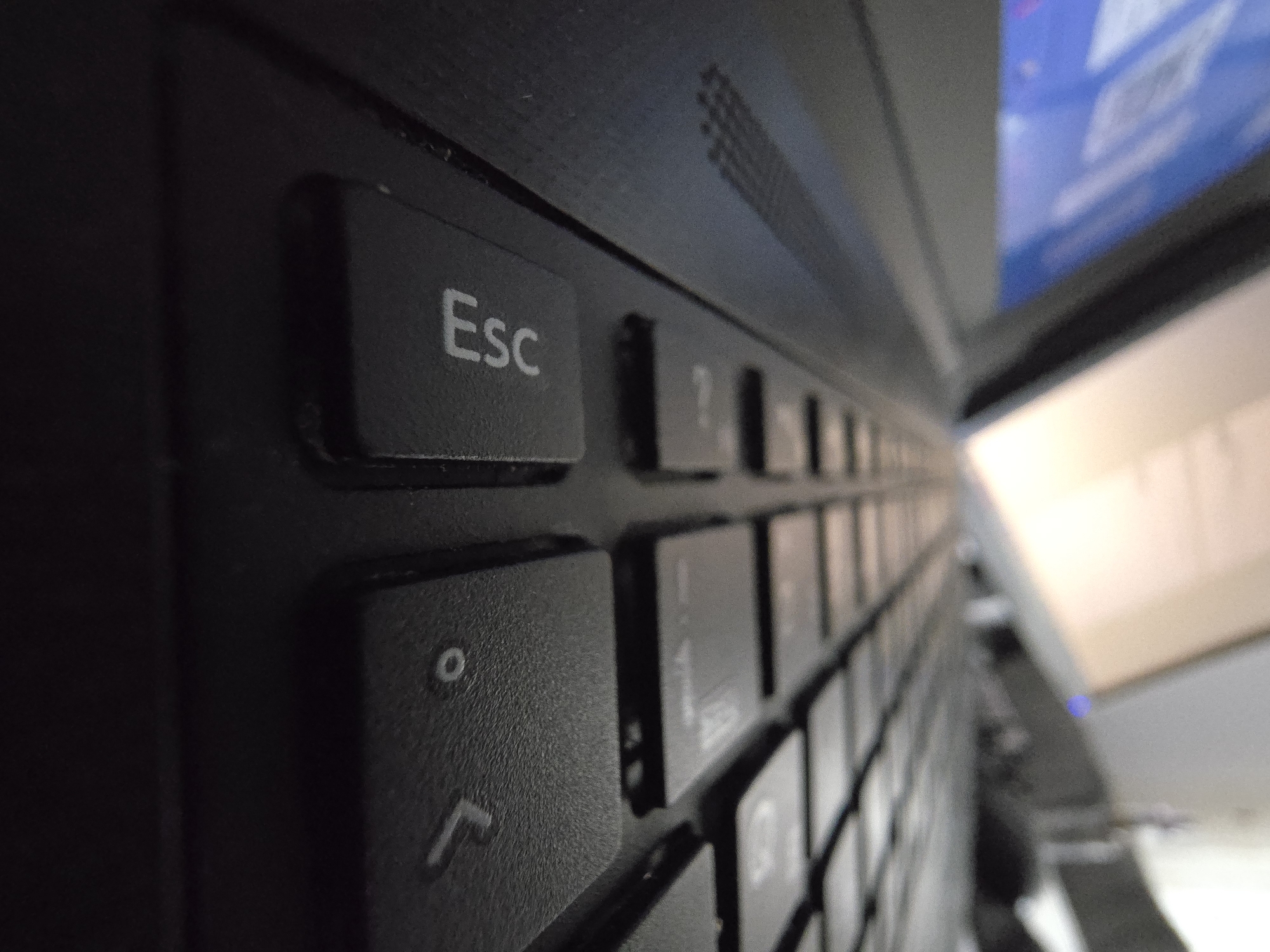 A close-up, angled shot focuses on a laptop keyboard. The "Esc" key is prominently visible on the left. The keys are black, and the lettering is light gray. The image has a shallow depth of field, with the keys receding into blur in the distance. The top right corner of the laptop and a glimpse of the screen can be seen. The environment is not discernible to provide a location.