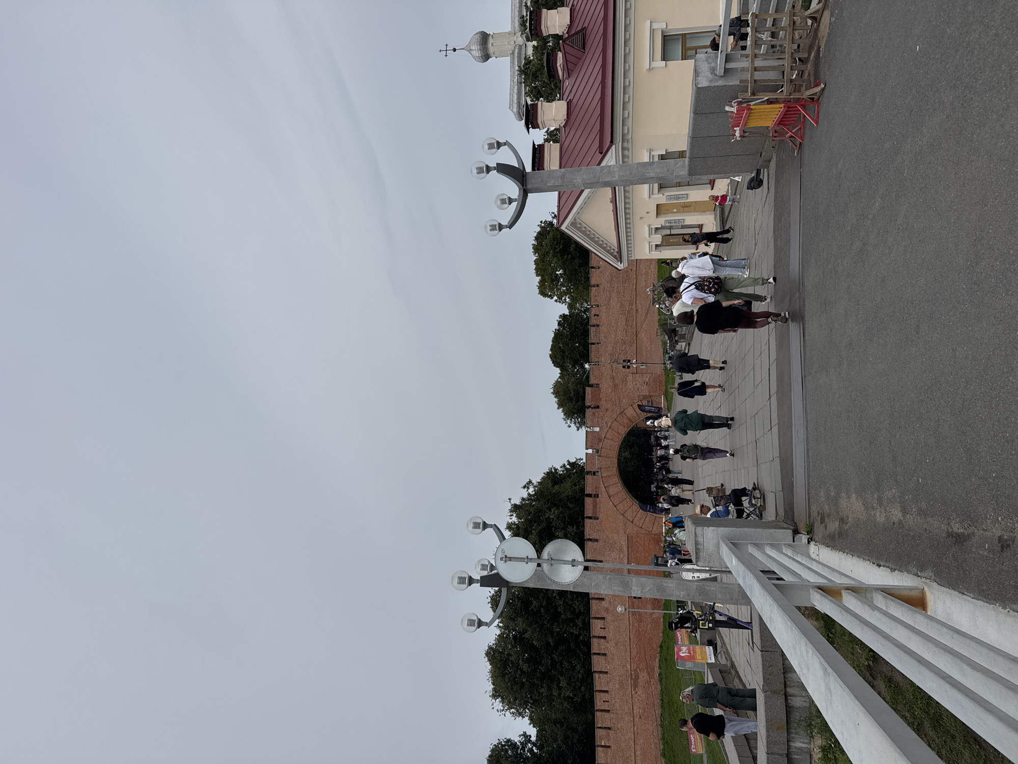 The image depicts a view of the Veliky Novgorod Kremlin in Russia. In the foreground, a paved walkway runs across a bridge, with a metal railing on the left. Several people are walking on the walkway. On the right side of the walkway, a section of a building with a red roof is visible, as well as a yellow facade with windows and doors. Beyond this, a large brick wall, characteristic of the Kremlin, stretches across the midground, with an arched gateway leading into the fortress. Tall trees line the area behind the wall. In the background, a domed building with a cross, likely a church or cathedral within the Kremlin, is visible against a cloudy sky. The overall scene captures a public space within a historical landmark.
